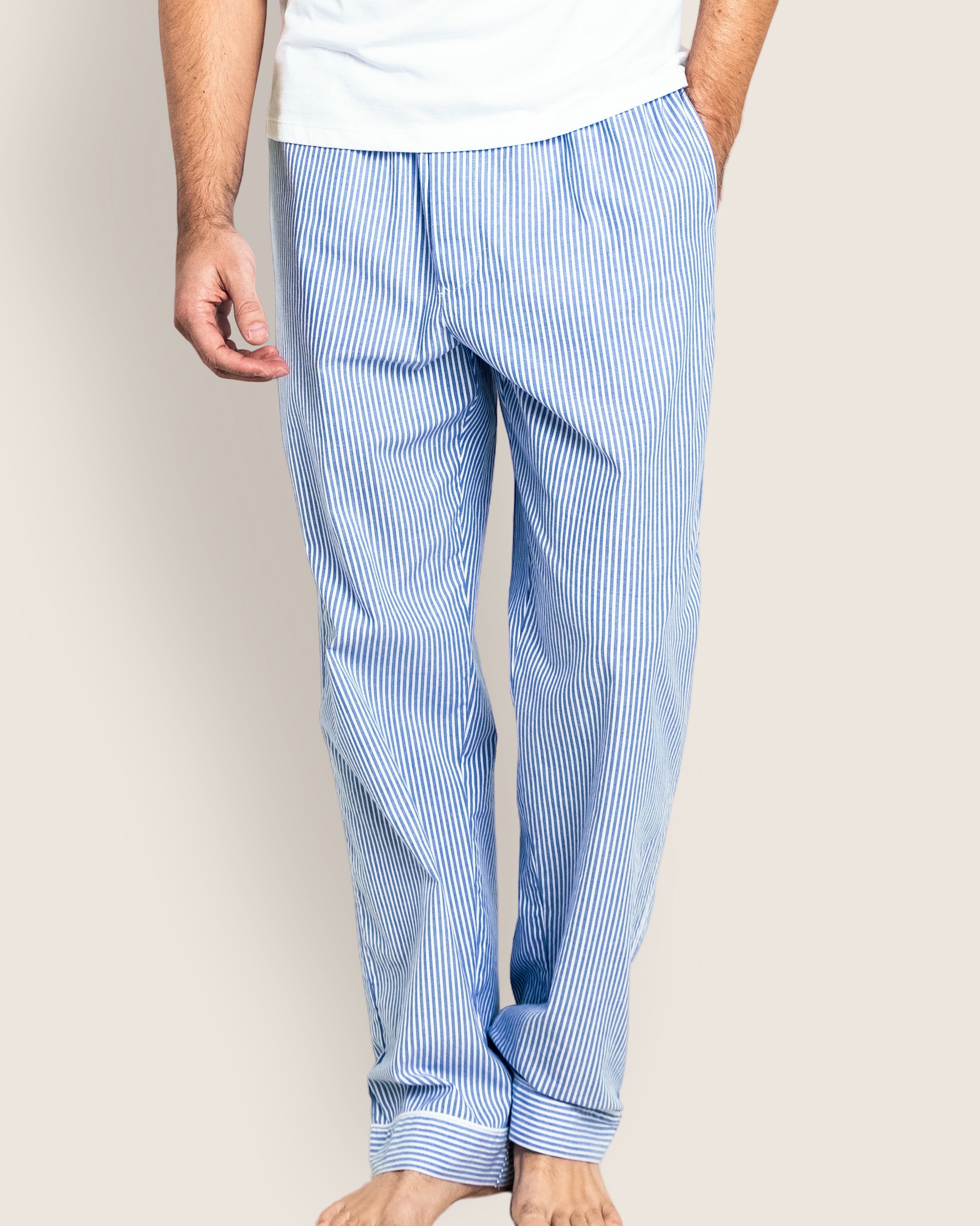 A person wearing Petite Plumes luxurious Mens Twill Pajama Pants in French Blue Seersucker stands barefoot against a neutral background, showcasing the light blue and white striped premium cotton pants along with a glimpse of the white shirt.