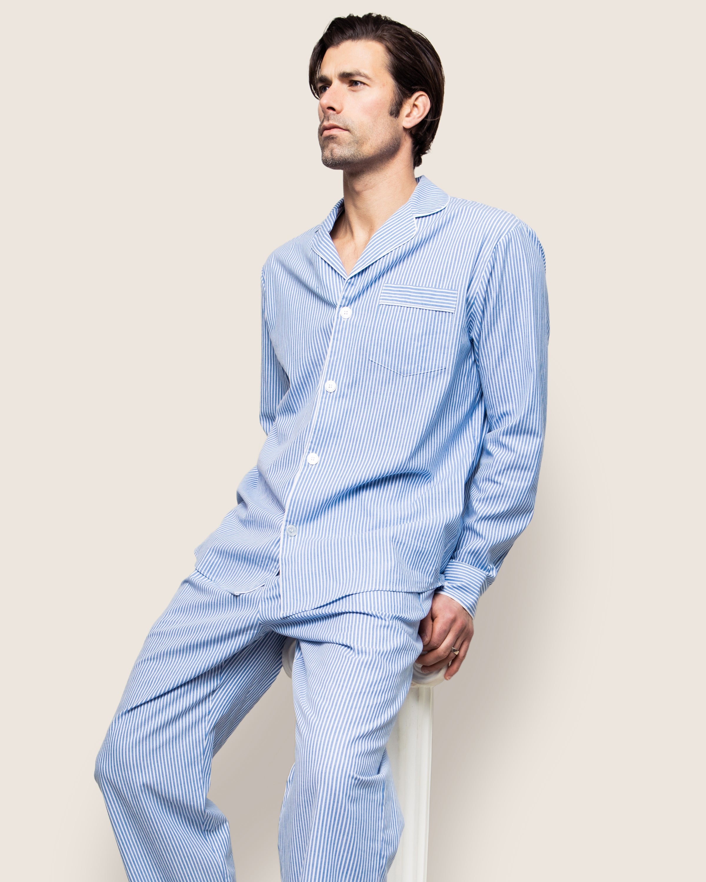 A person in a Petite Plume Mens Twill Pajama Set in French Blue Seersucker sits on a stool against a simple backdrop. Made from premium cotton, they have short, dark hair and look to the side with a serene expression.