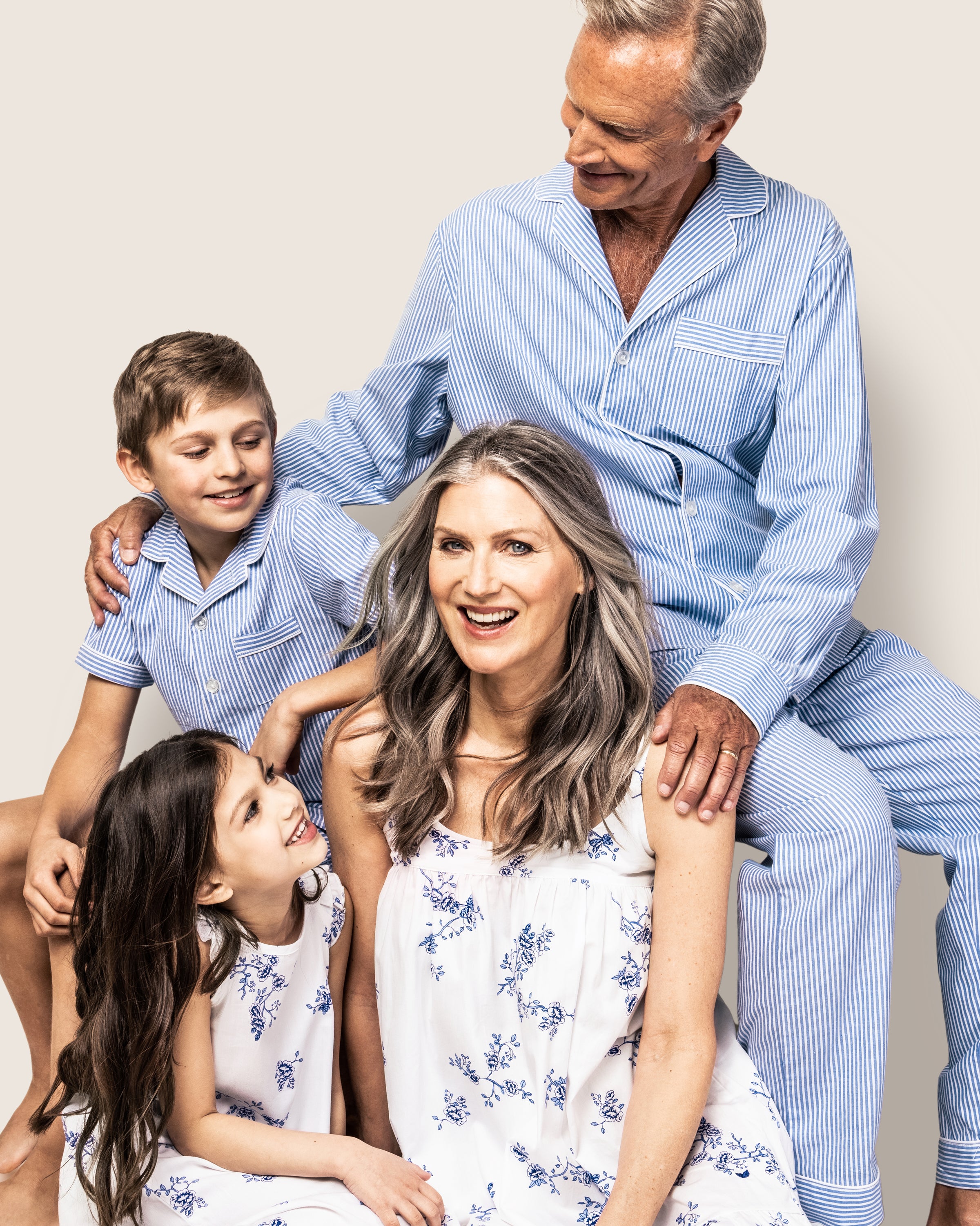 A family in matching Petite Plume blue and white pajamas gathers cozily. An older man in the Mens Twill Pajama Set in French Blue Seersucker sits with a boy, while a woman sits with a smiling young girl, adding joy to their gathering.