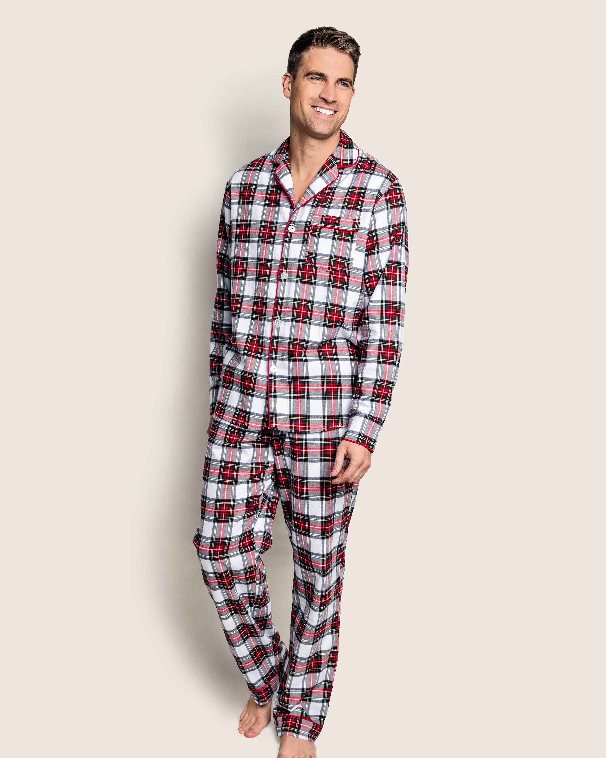 A man smiles while barefoot, clad in a Petite Plume Mens Brushed Cotton Pajama Set in Balmoral Tartan. The red, white, and black plaid ensemble includes a button-up long-sleeve top and matching pants made from high-quality cotton. He poses against a plain, light-colored background.