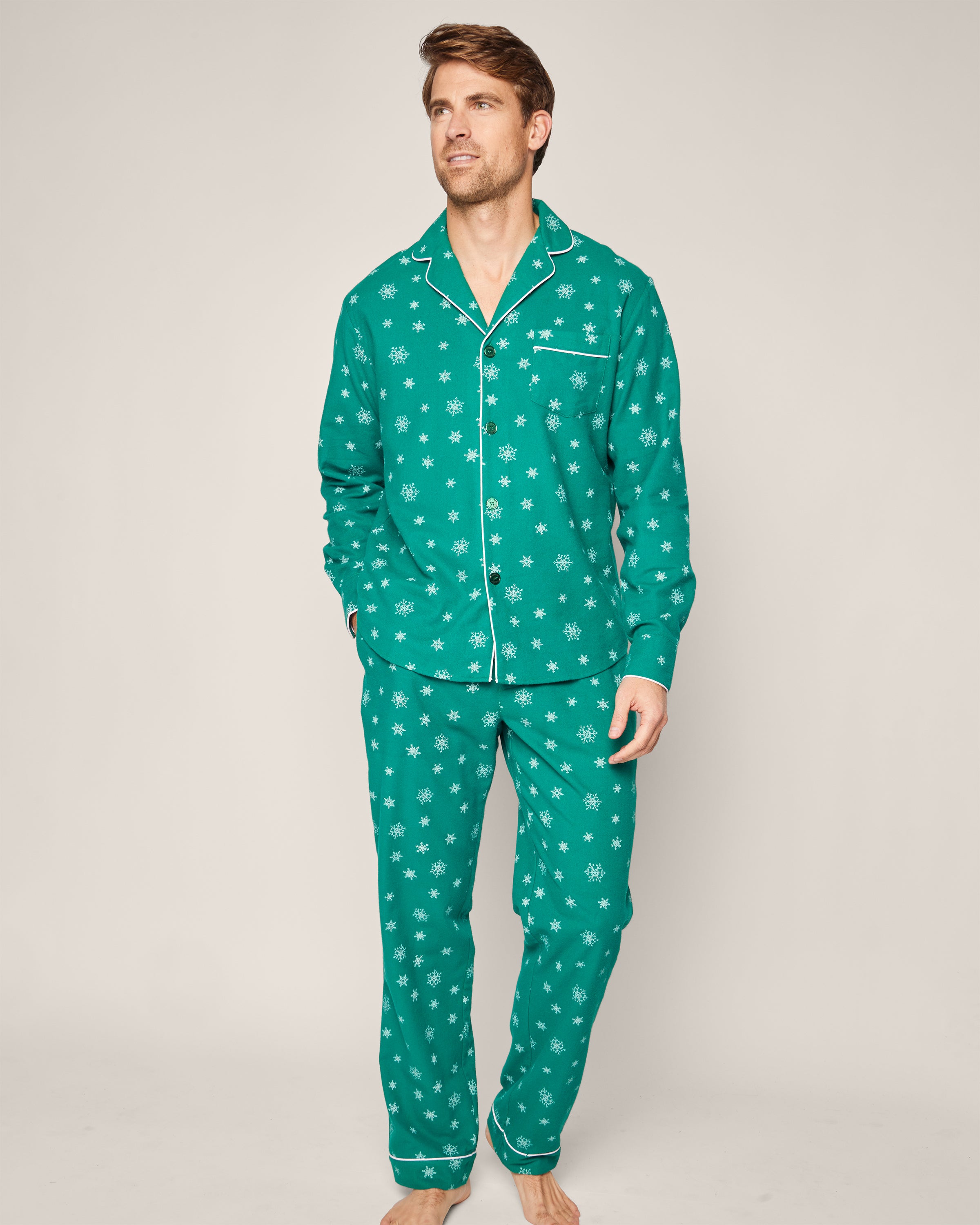 A person stands barefoot against a plain background, wearing Petite Plumes Mens Flannel Pajama Set in Emerald Wonderland. This set is made from premium cotton, featuring an emerald green color with white snowflakes and includes a classic collared, long-sleeved top.
