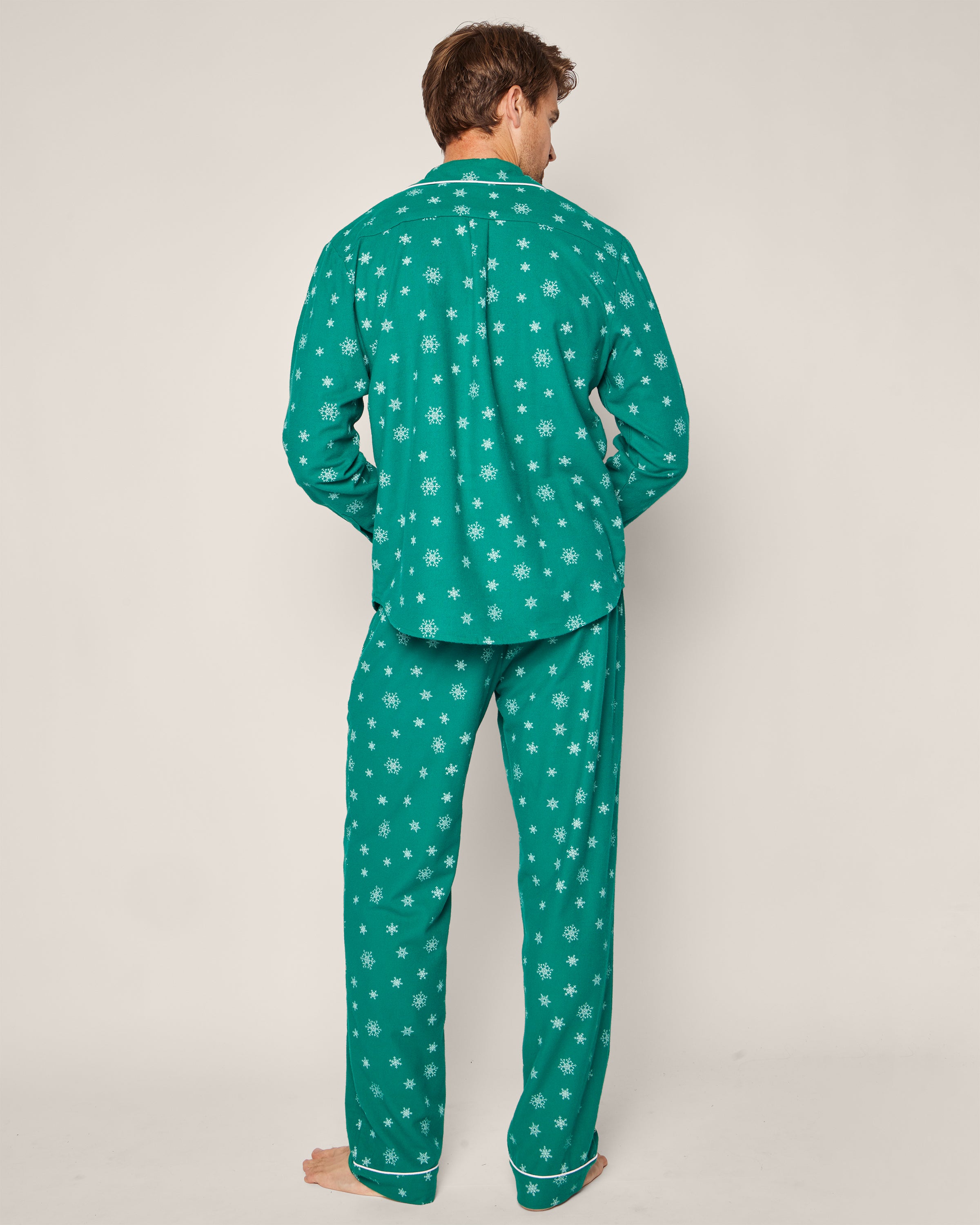 A person is seen from behind wearing Petite Plumes Mens Flannel Pajama Set in Emerald Wonderland, featuring white snowflakes. Crafted from premium cotton, it includes a long-sleeve shirt and pants. The backdrop is plain and light-colored.