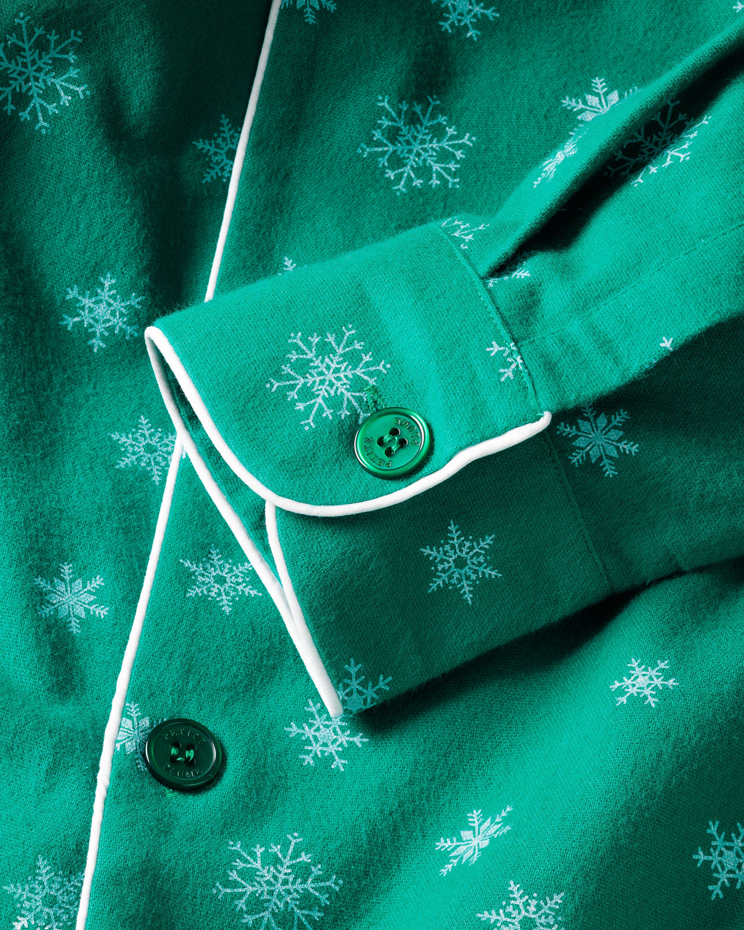 A close-up of the Mens Flannel Pajama Set in Emerald Wonderland from Petite Plume displays an emerald green flannel with white snowflakes, a buttoned cuff with white piping, and a green button, embodying timeless classic pajama charm.