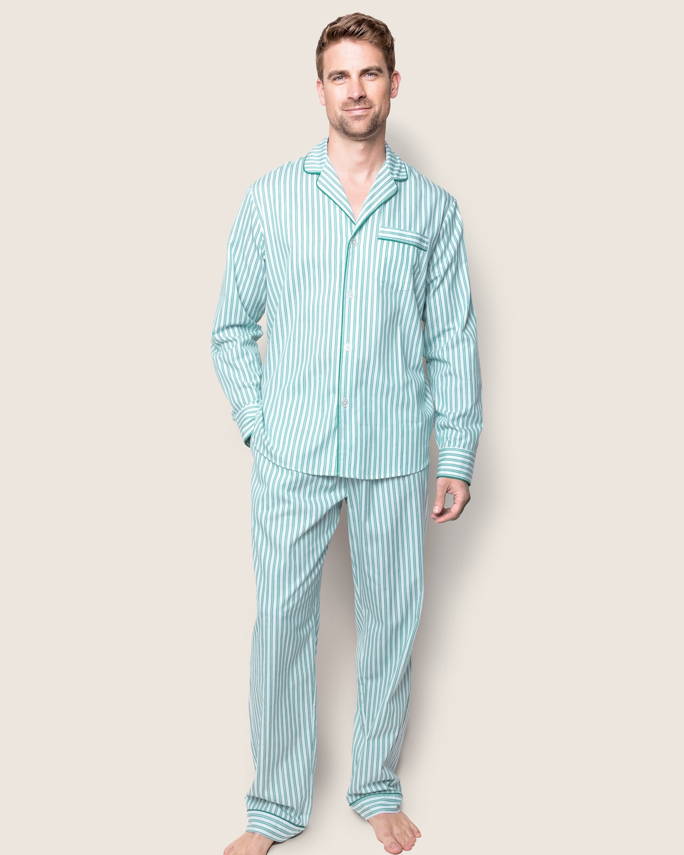 A man stands against a beige background wearing Petite Plumes Mens Twill Pajama Set in Emerald Ticking. The set features a button-up top with a pocket and matching pants in a subtle French ticking pattern. He is barefoot and smiles slightly at the camera.