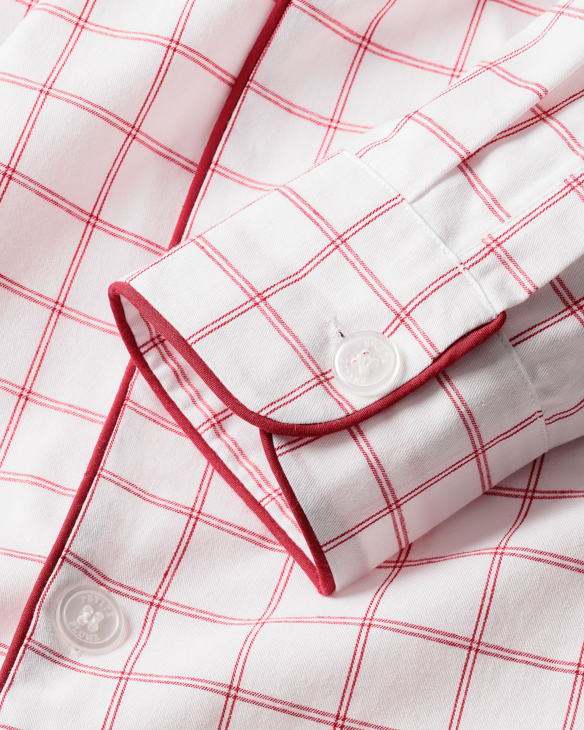 Close-up of Petite Plumes Mens Twill Pajama Set in Garnet Tattersall, featuring a white shirt with a garnet-red grid pattern. Made from 100% cotton twill, it includes a buttoned cuff with red piping for an elegant touch.