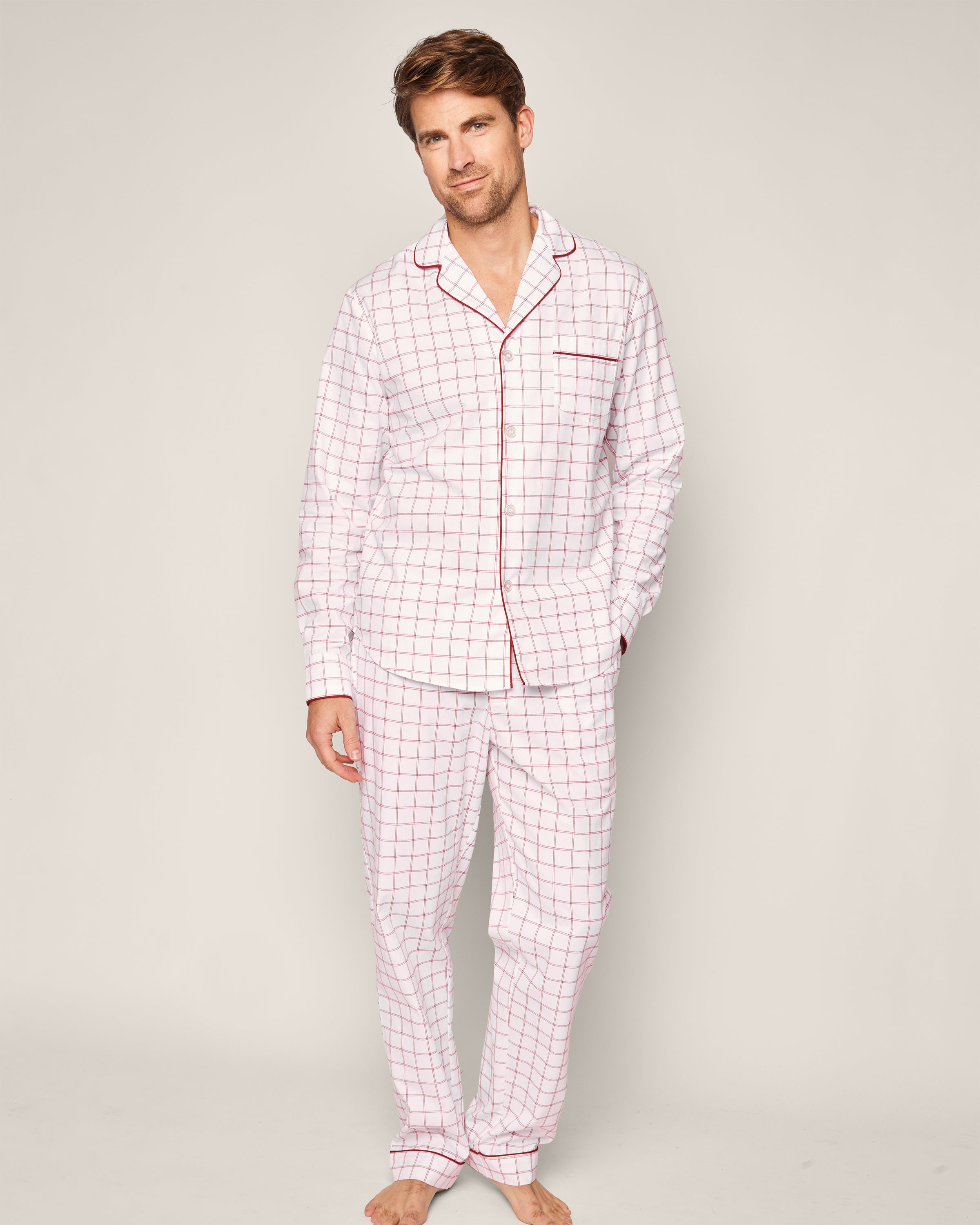 A person with short hair and a neutral expression stands barefoot against a plain background wearing the Mens Twill Pajama Set in Garnet Tattersall by Petite Plume. Crafted from 100% cotton twill, the set includes a button-up top and long pants.