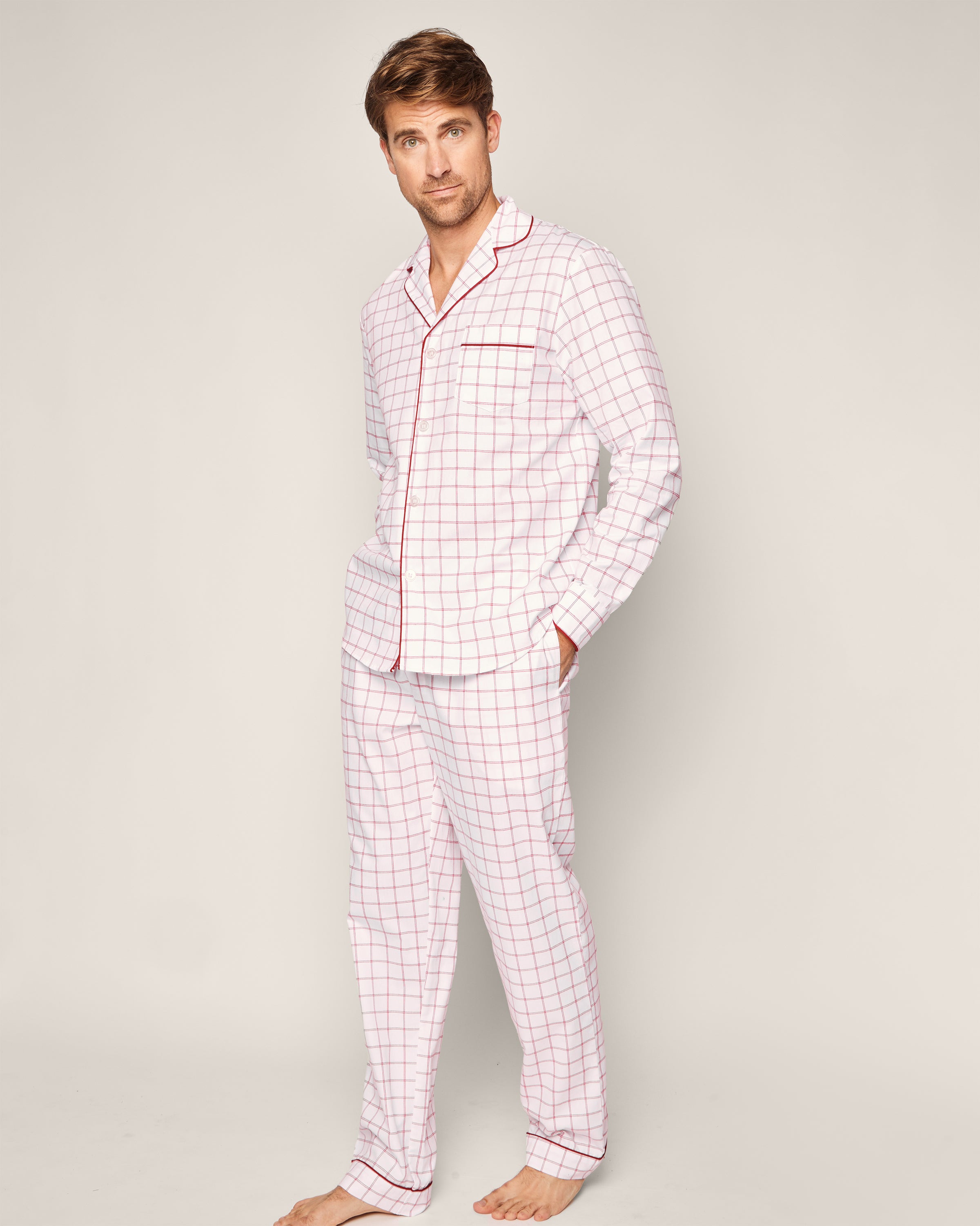 A man stands barefoot in a relaxed pose wearing Petite Plumes Mens Twill Pajama Set in Garnet Tattersall, made from 100% cotton. His hands rest casually in his pockets against a neutral background, epitomizing comfort and style.