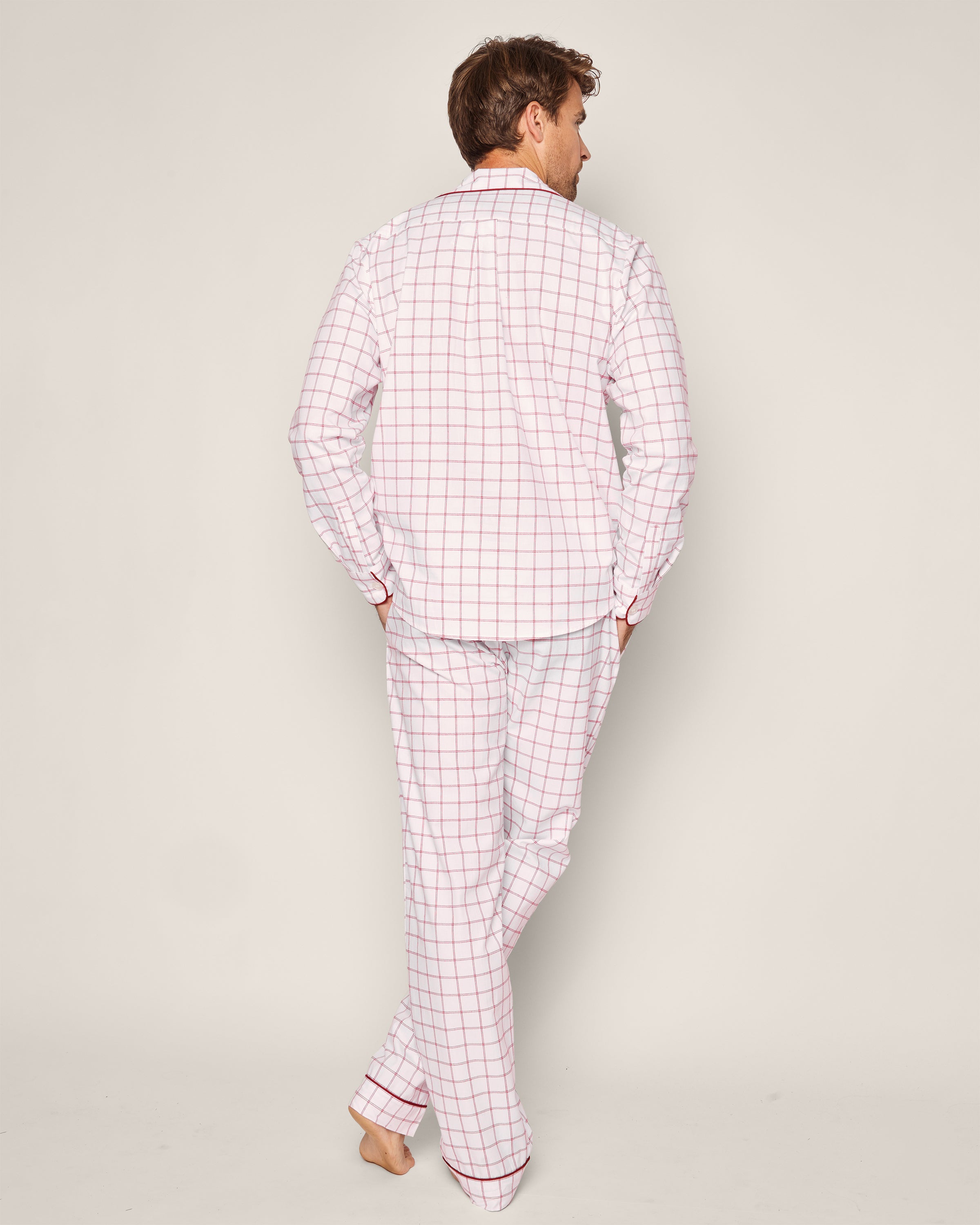 A person stands barefoot, back to the camera, wearing the Mens Twill Pajama Set in Garnet Tattersall by Petite Plume, featuring a matching long-sleeved shirt and pants with a white and pink grid on garnet. The background is plain white.