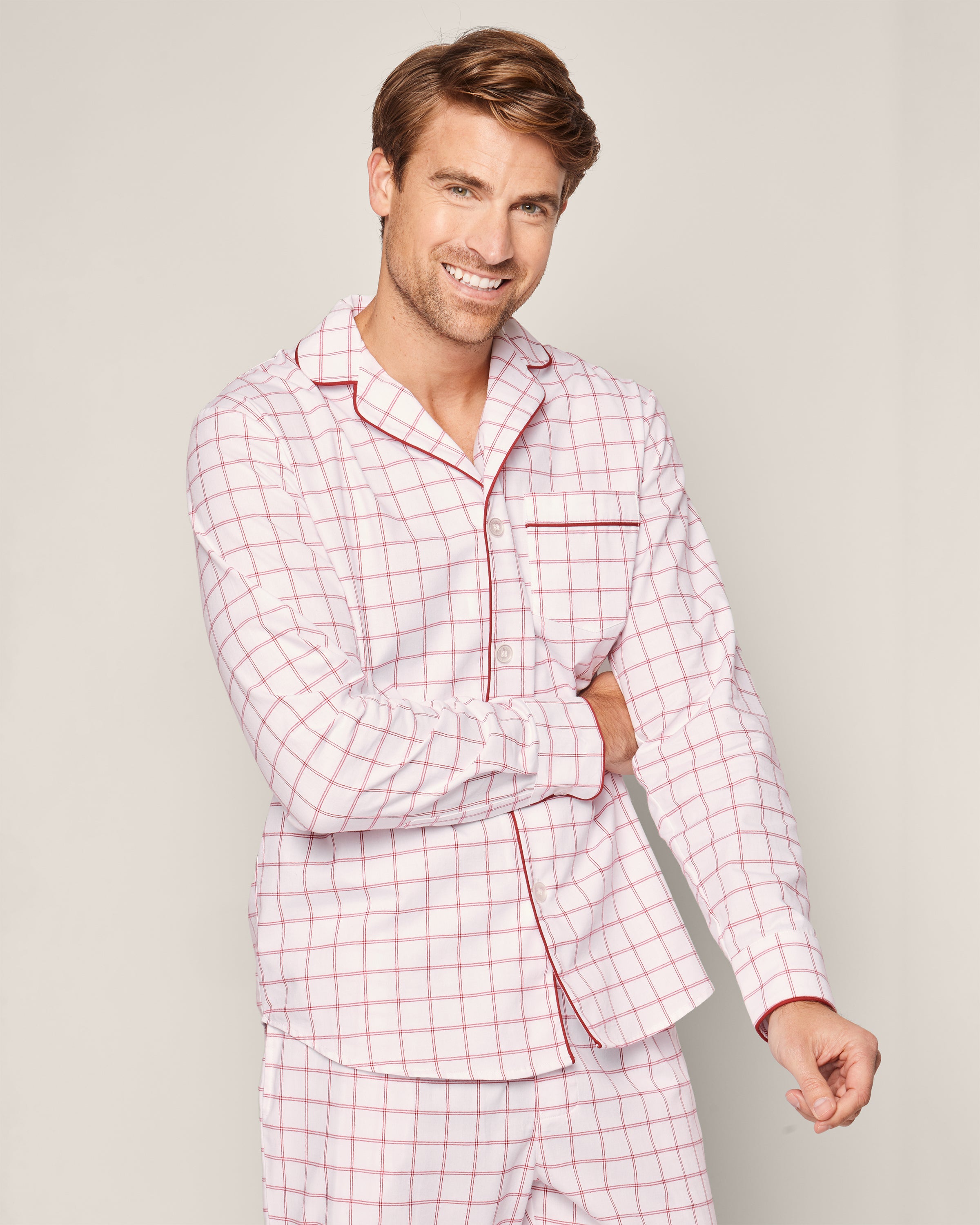 A person smiling, wearing the Mens Twill Pajama Set in Garnet Tattersall by Petite Plume. Made of 100% cotton twill, this set has a classic garnet-red checkered design with red piping, a collar, and a chest pocket. The plain light background adds to its cozy appeal.