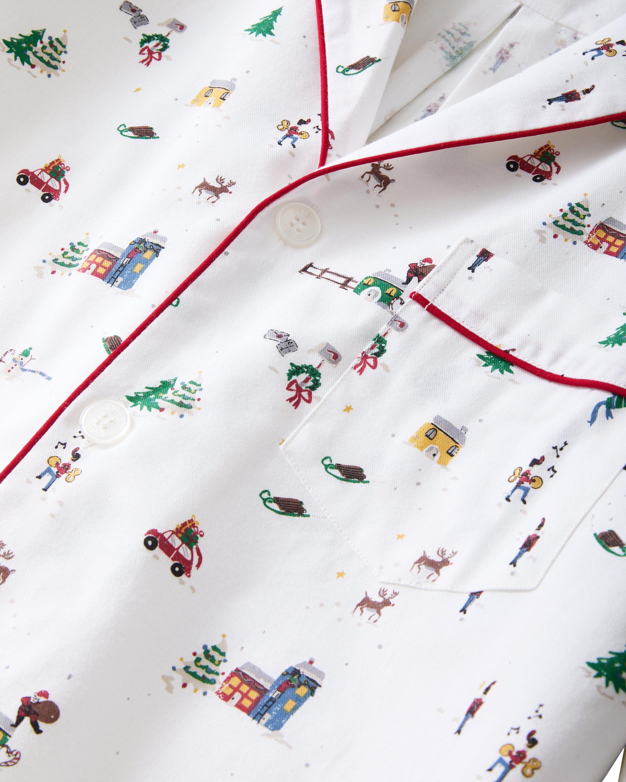 A man stands barefoot against a plain background, wearing the Men's Twill Pajama Set in Holiday Town, featuring a small, colorful print, red piping, a collared button-up top, and long pants.