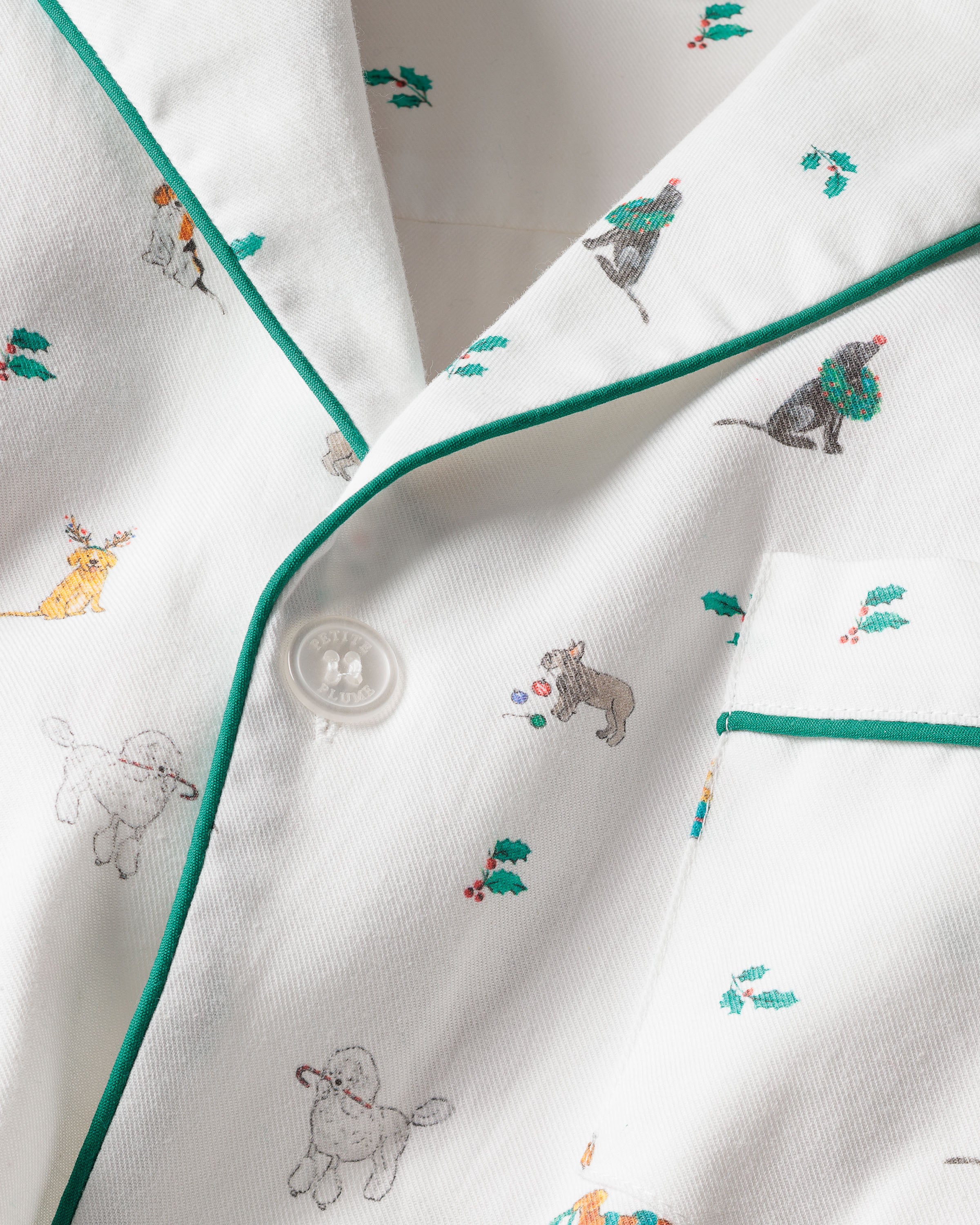 Close-up of Petite Plumes Mens Twill Pajama Set in Jingle Paws: a white cotton twill top featuring playful canine designs with green piping, some wearing festive hats and scarves, surrounded by scattered green and red holly leaves and berries, ideal for holiday sleepwear.