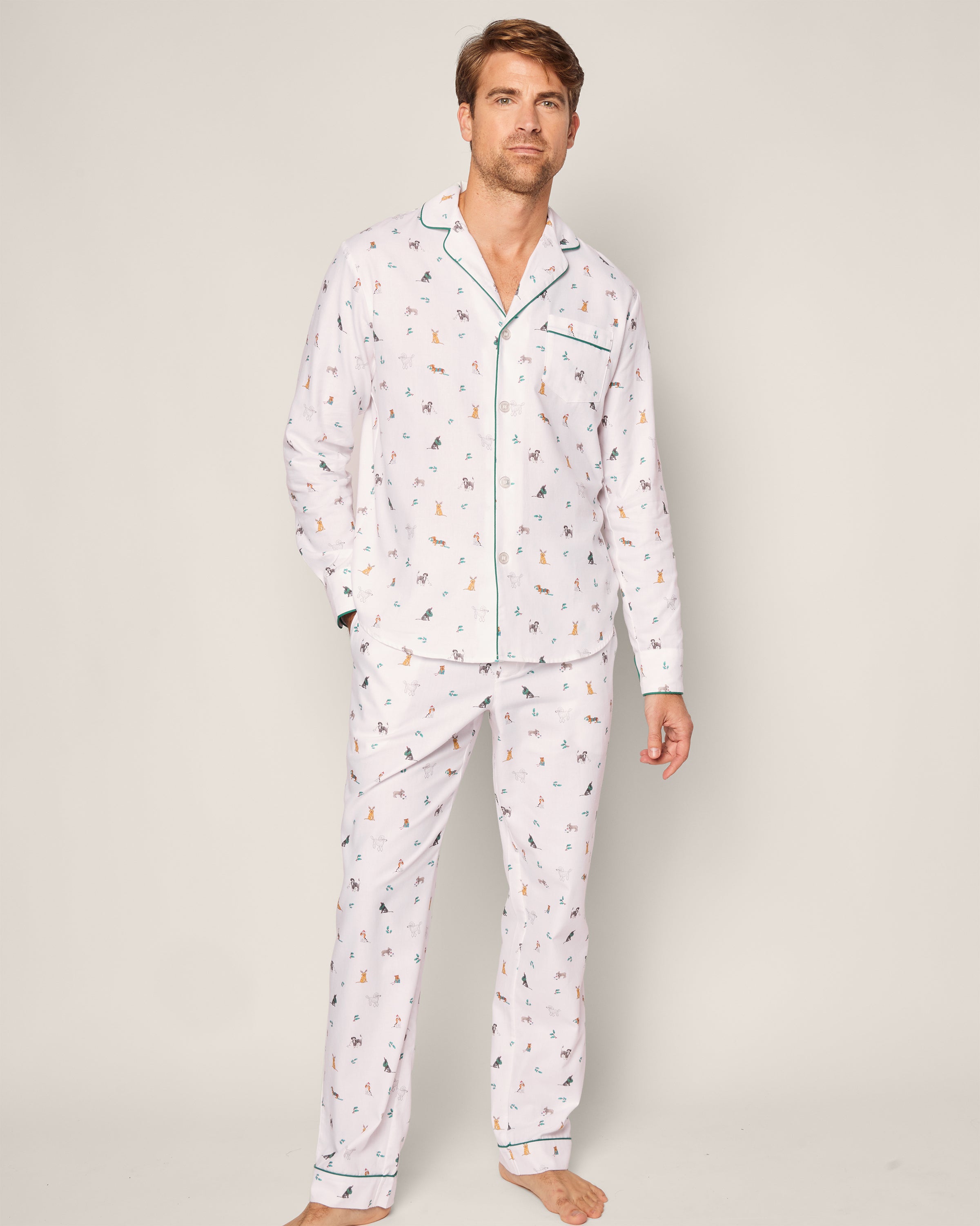 A man models the Mens Twill Pajama Set in Jingle Paws by Petite Plume, featuring animal-patterned white pajamas made from premium cotton. The set includes a button-up shirt with a collar, long pants, and dark piping. Hes barefoot against a plain background.