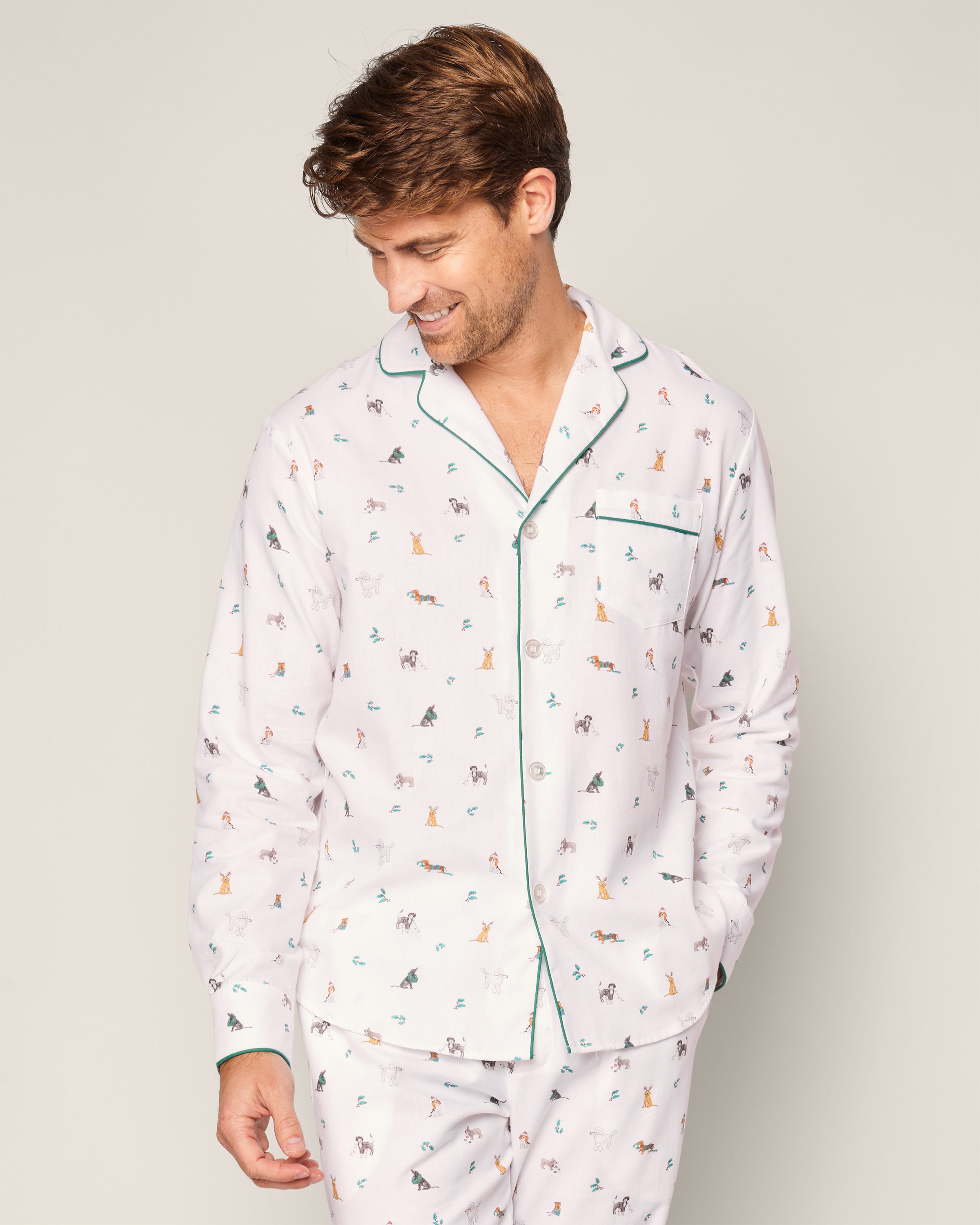 A man in Petite Plumes Mens Twill Pajama Set in Jingle Paws, featuring a white fabric adorned with colorful animal prints and green-trimmed collar and pocket, looks downward with a slight smile against a light grey background.