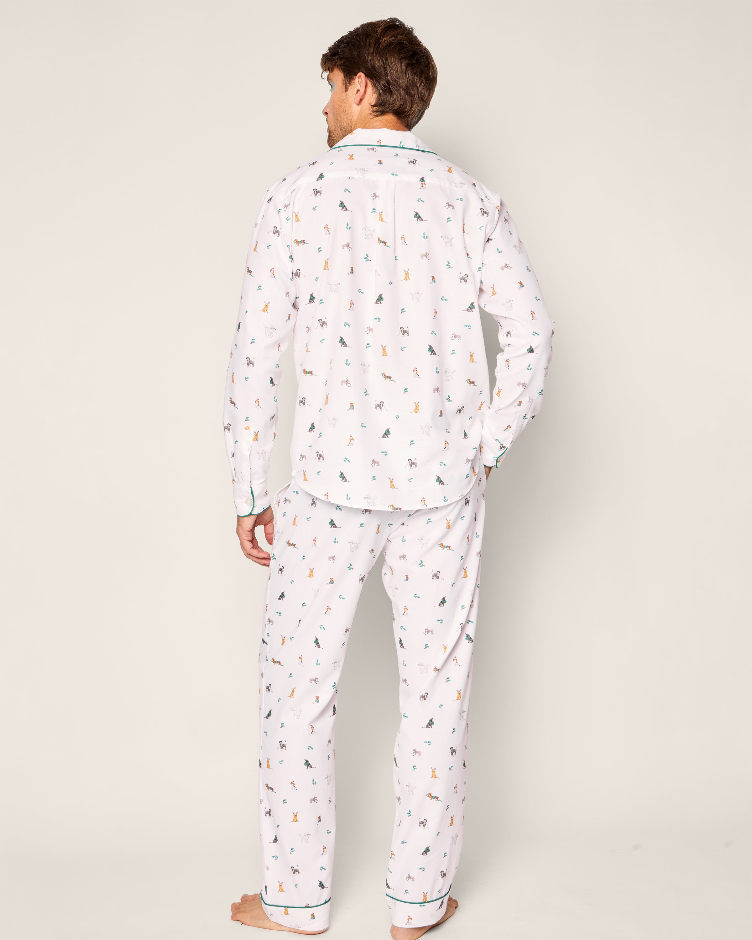 A person stands barefoot in Petite Plumes Mens Twill Pajama Set in Jingle Paws, featuring a holiday design with tiny multicolored prints. The relaxed-fit, long-sleeved set is made from premium cotton against a plain light-colored backdrop.