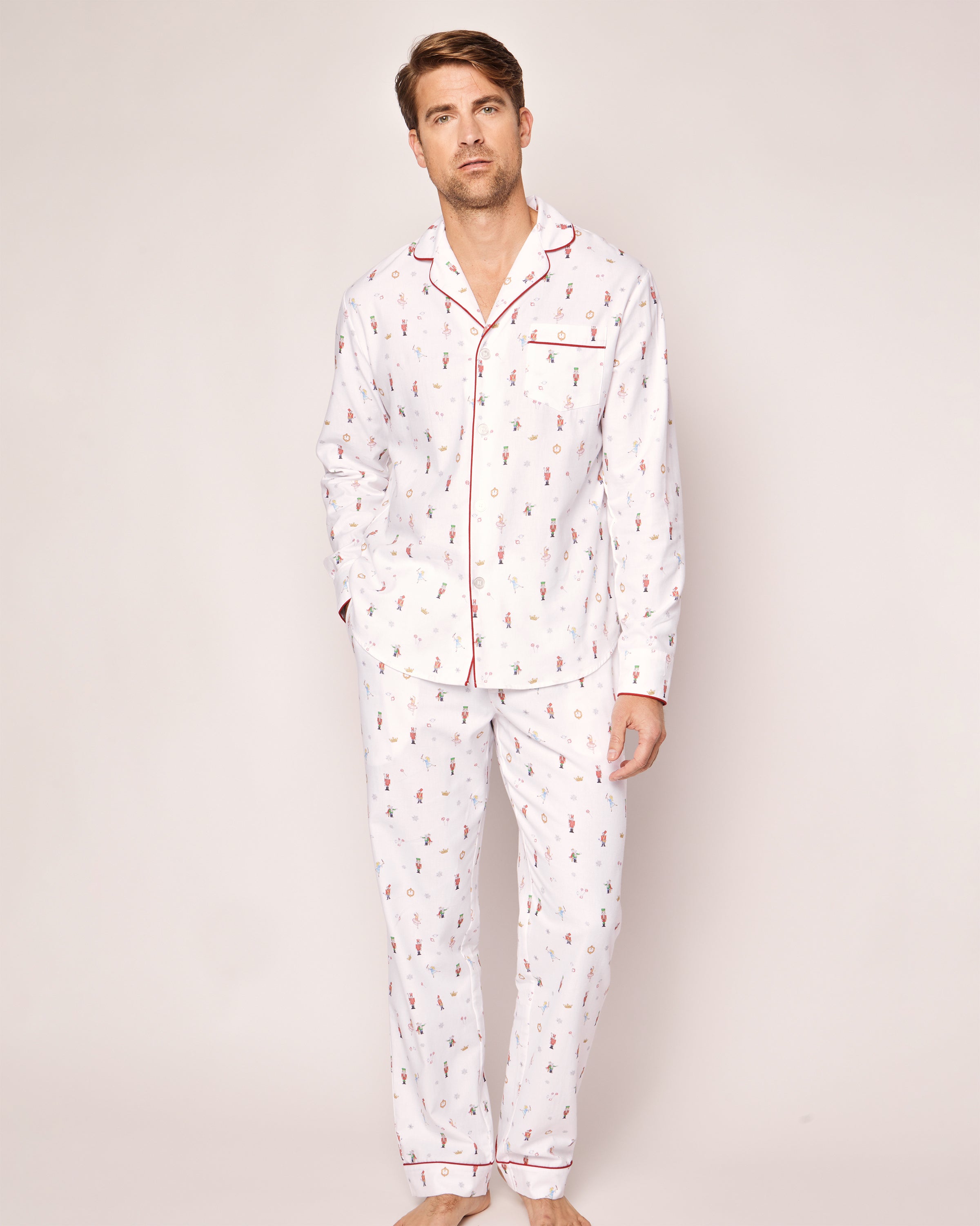 A person stands against a plain background in Petite Plumes luxurious Mens Twill Pajama Set, A Night at the Nutcracker. Featuring small printed patterns, red piping, and a button-up top with a pocket, these pajamas echo classic Nutcracker style.