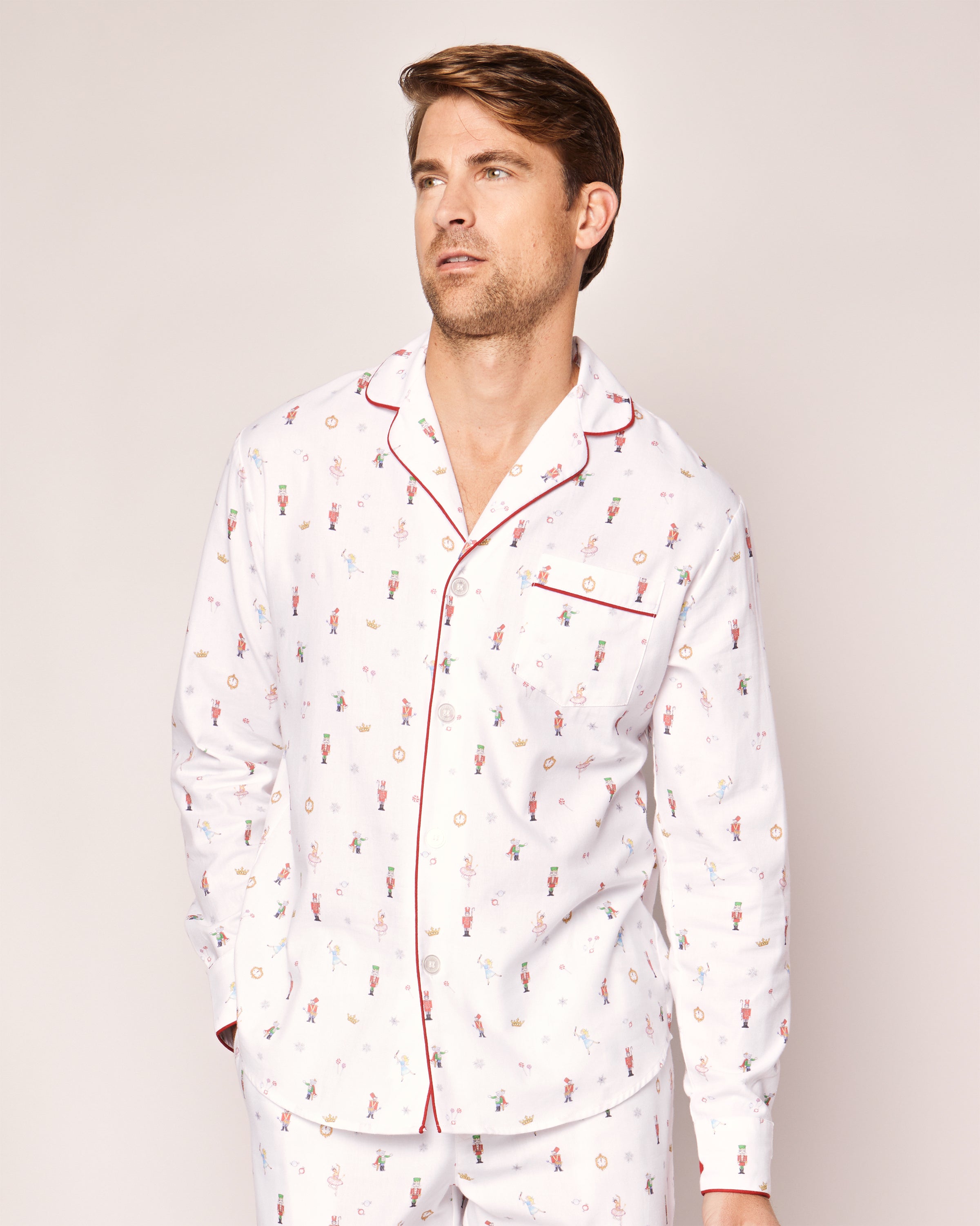 A man wearing Petite Plumes Mens Twill Pajama Set in A Night at the Nutcracker, featuring pajamas with colorful prints and red piping, stands with hands in pockets, gazing sideways against a light-colored background.