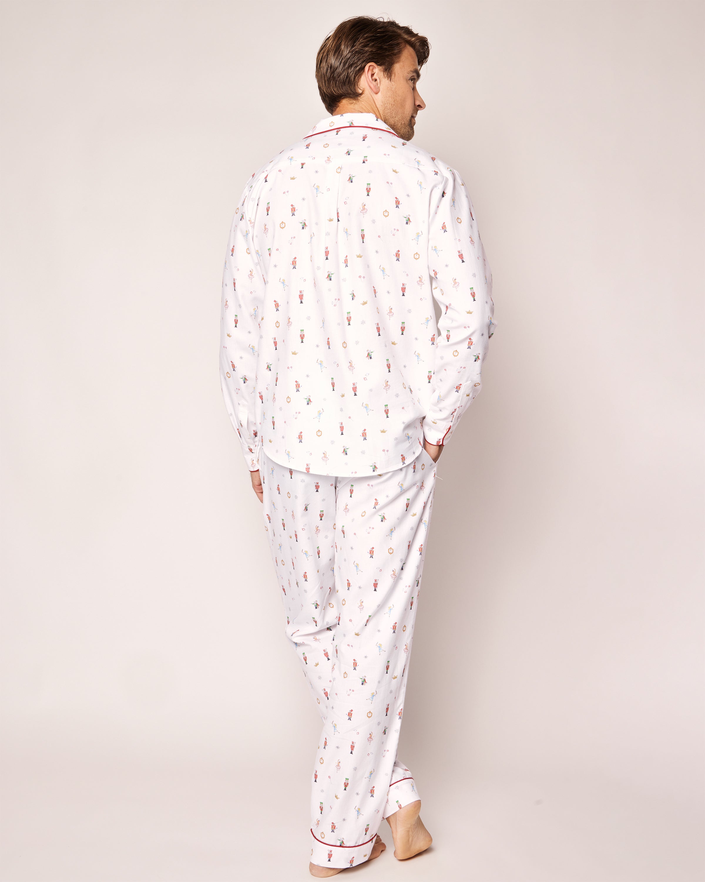 Barefoot and facing away, the individual wears Petite Plumes Mens Twill Pajama Set in A Night at the Nutcracker, featuring luxury sleepwear with a small red pattern and elegant red piping. These pajamas add sophistication against a plain, neutral background.