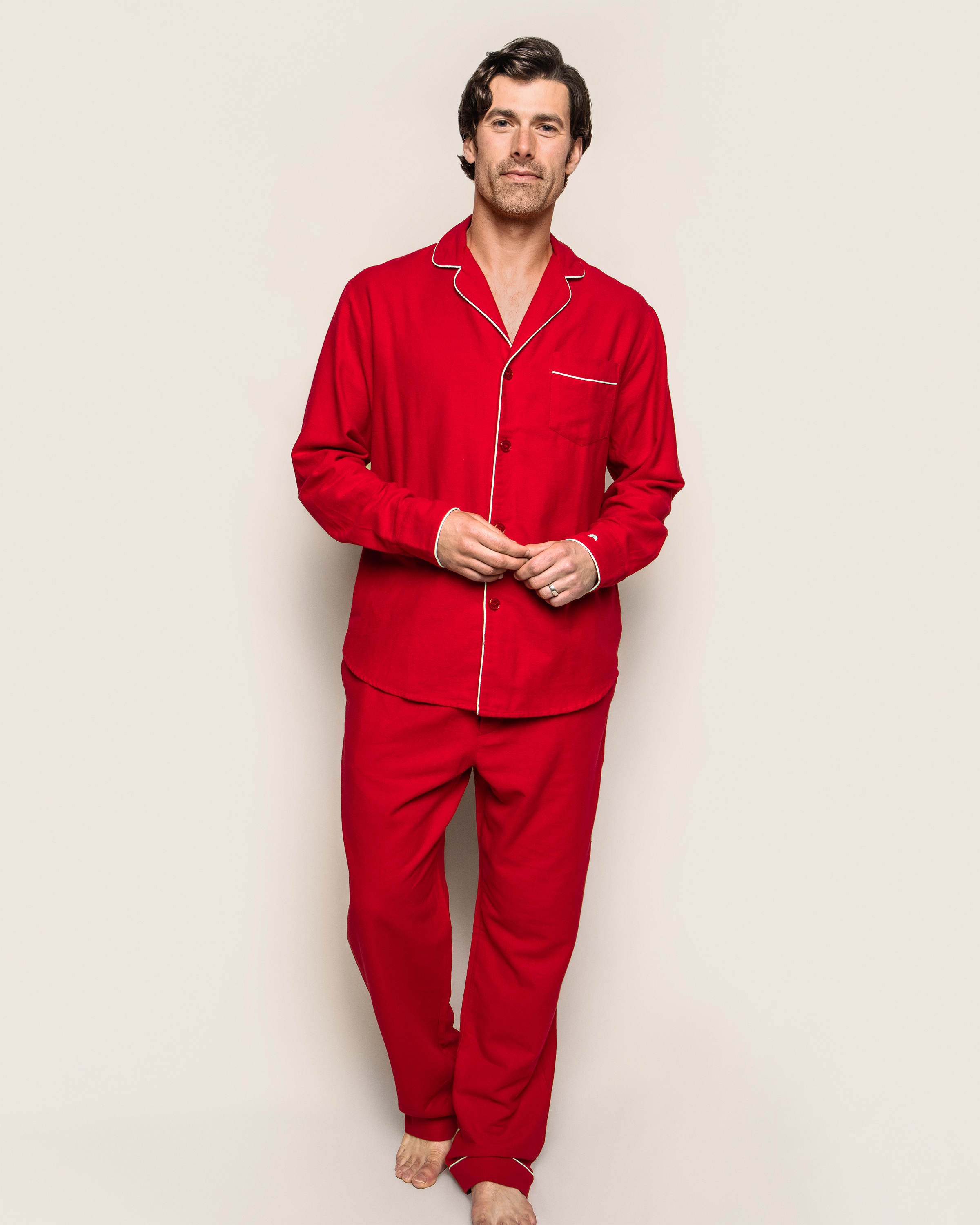 Against a plain backdrop, a person wears Petite Plumes Mens Flannel Pajama Set in red with white piping. Their dark hair and neutral expression highlight the elegance of the 100% cotton attire. Barefoot, they exude a relaxed and sophisticated vibe.