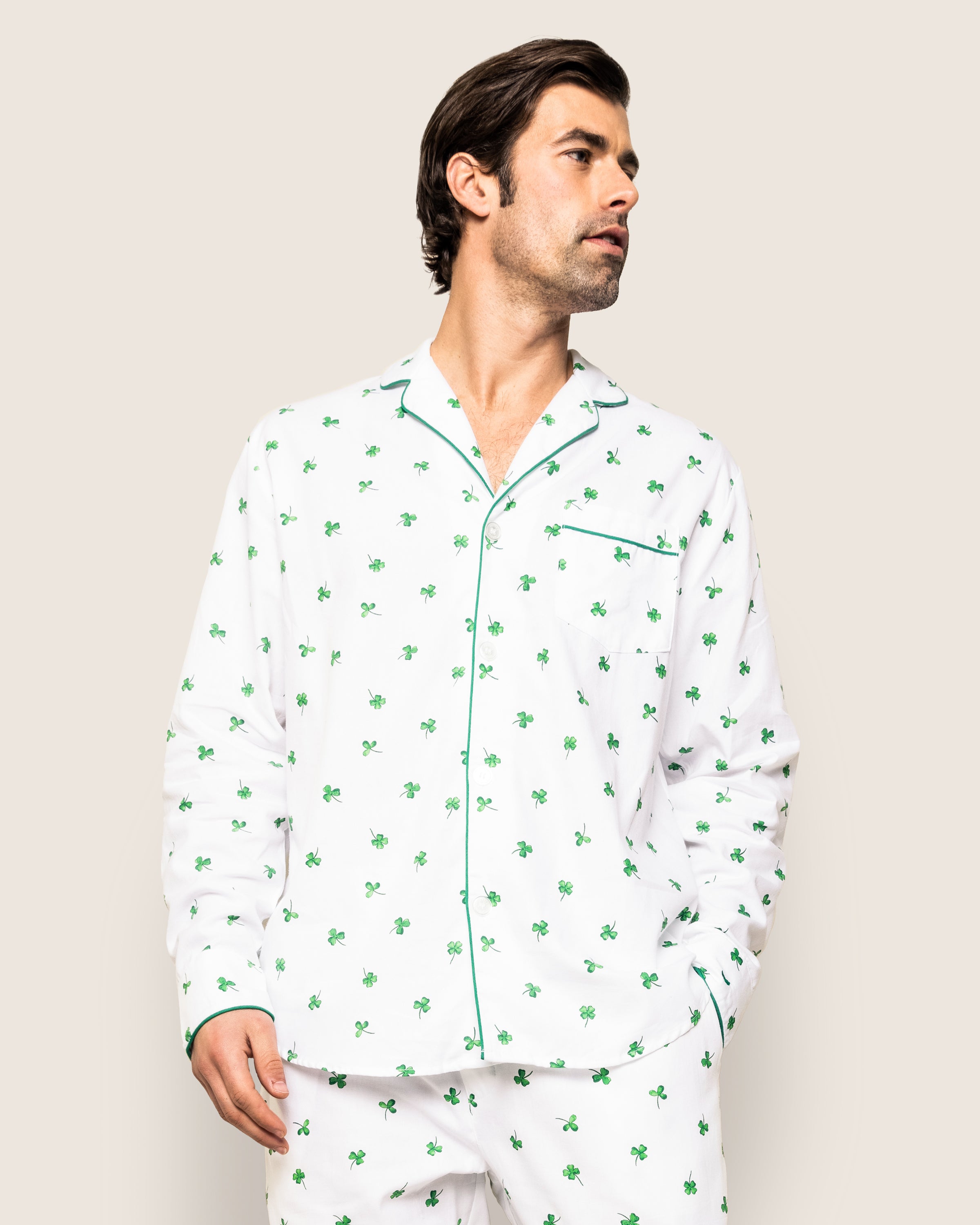 A man models the Mens Twill Pajama Set in Shamrocks by Petite Plume, showcasing white fabric with small green palm trees. Standing against a plain backdrop, he gazes left with relaxed hands, highlighting the elegant green piping along the edges.