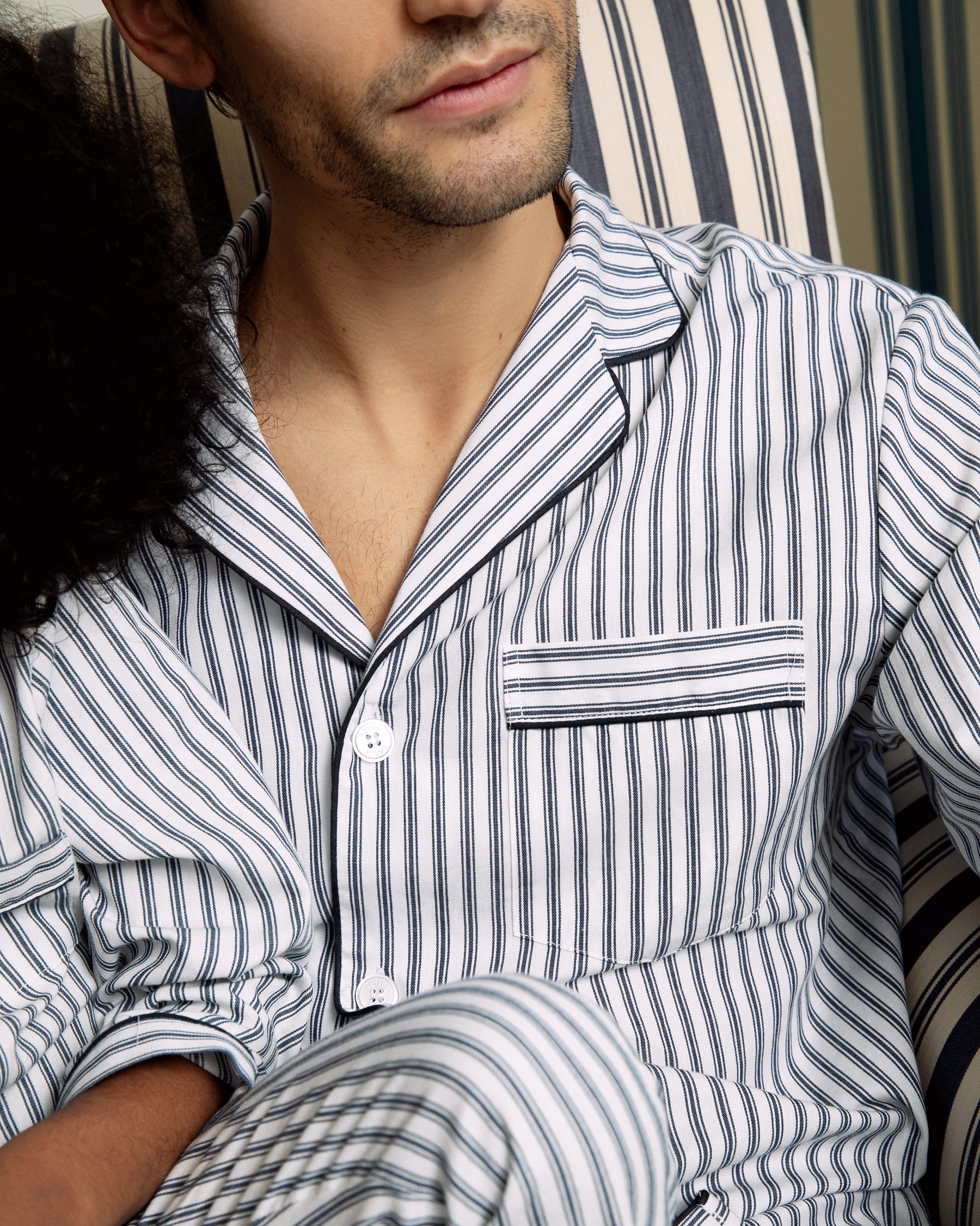 A person is seated on a chair with a matching design, wearing Petite Plumes Mens Twill Pajama Set in Navy French Ticking. The image highlights the pajamas button and pocket details, showcasing the finest quality cotton in its construction.