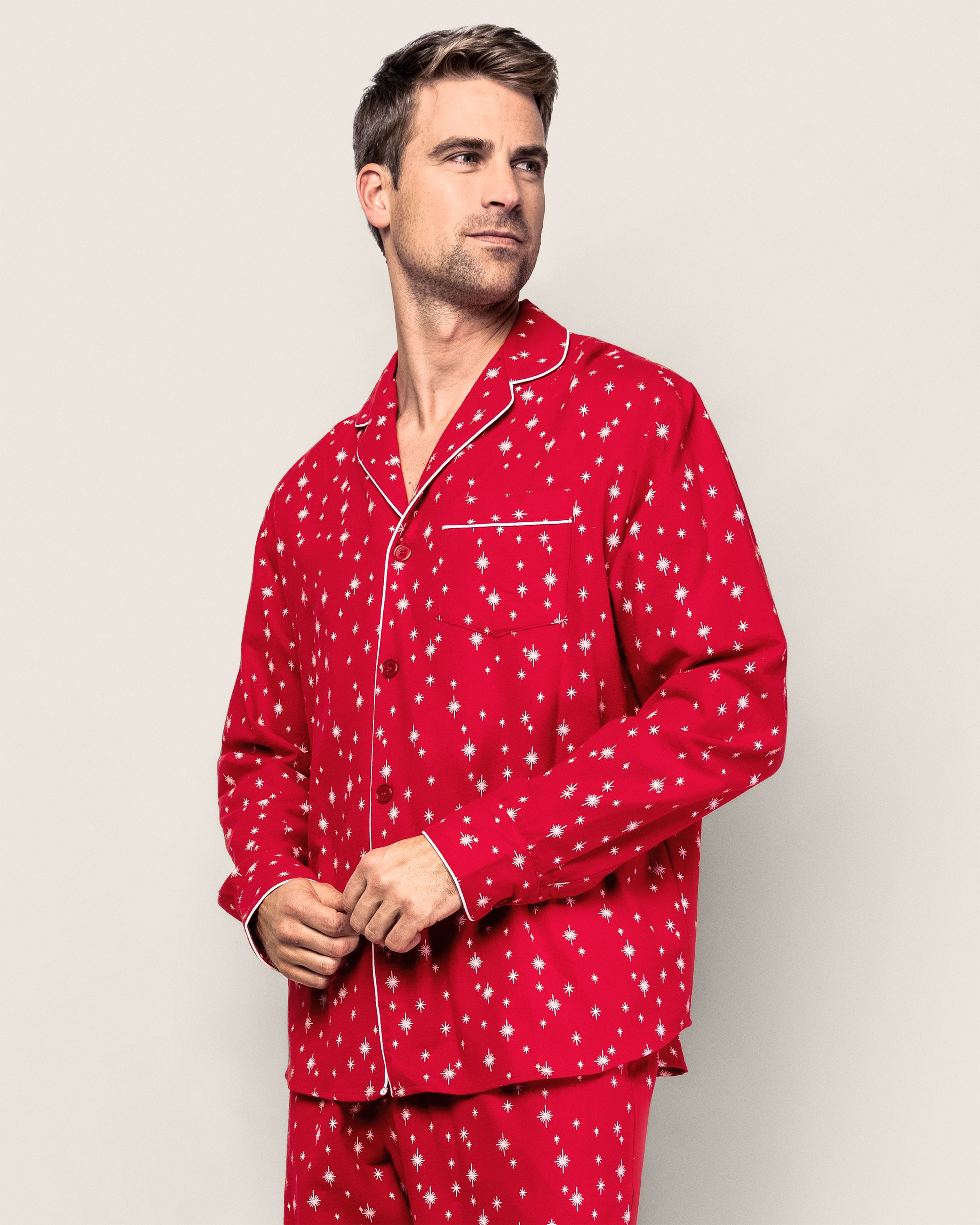 Wearing Petite Plumes Mens Flannel Pajama Set in Starry Night, a man stands against a plain background. The 100% cotton attire boasts red fabric with white star patterns, long sleeves, and a top pocket. He looks to the side, exuding comfort and style.