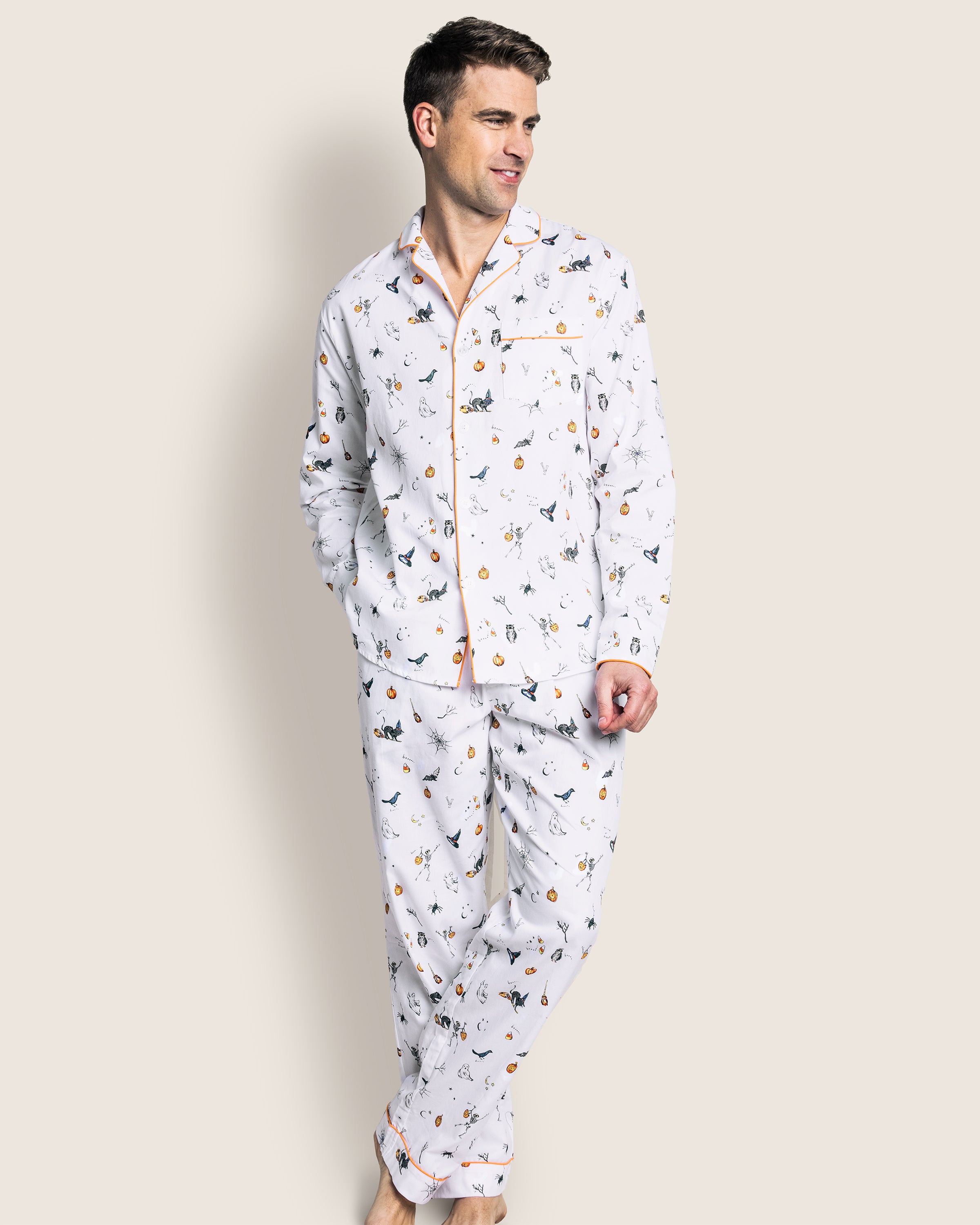 A man models Petite Plumes Mens Twill Pajama Set in Trick or Treat, featuring a playful bird design and orange trim, crafted from premium cotton for comfort and style. He stands against a plain background, hands in pockets and barefoot.