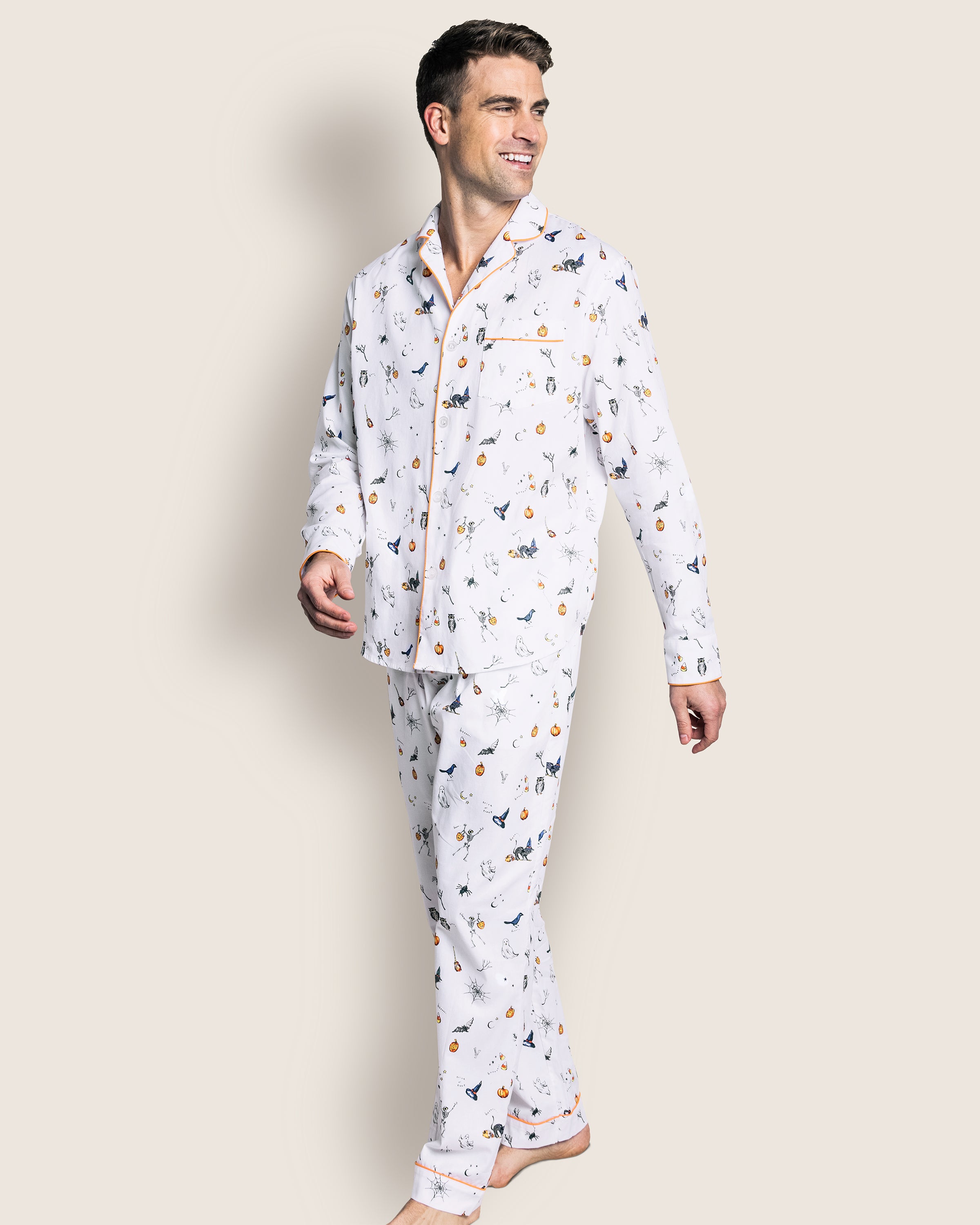 A man wearing Petite Plumes Mens Twill Pajama Set in Trick or Treat, featuring high-quality cotton with a colorful print, smiles and looks to the side. His pajamas include a chest pocket and piping details against a plain background.