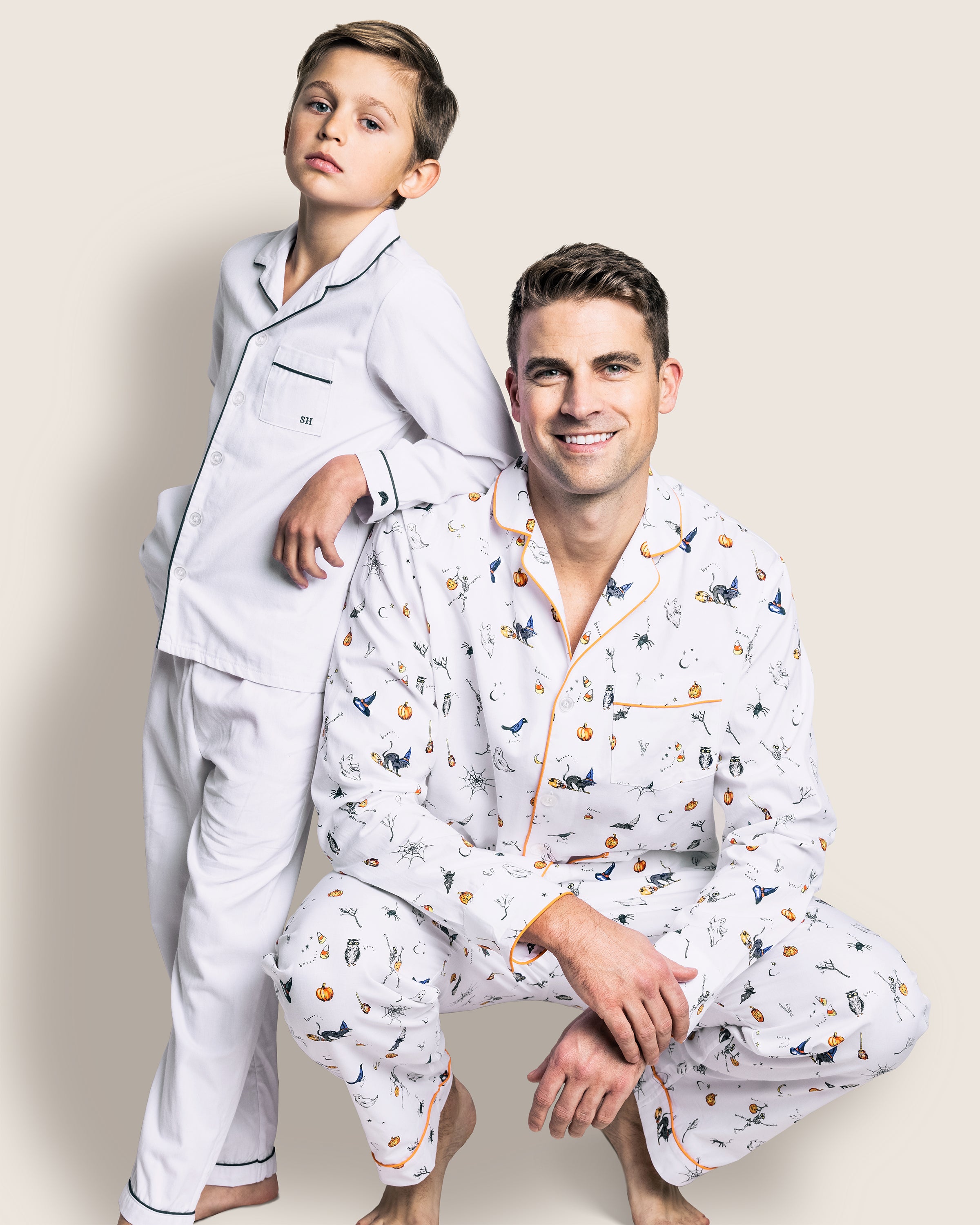 A man and boy in matching Halloween pajamas from Petite Plume pose together; the boy, in white pajamas with black piping, stands with his hand on the mans shoulder, while the man crouches smiling in a Mens Twill Pajama Set featuring a colorful Trick or Treat print.