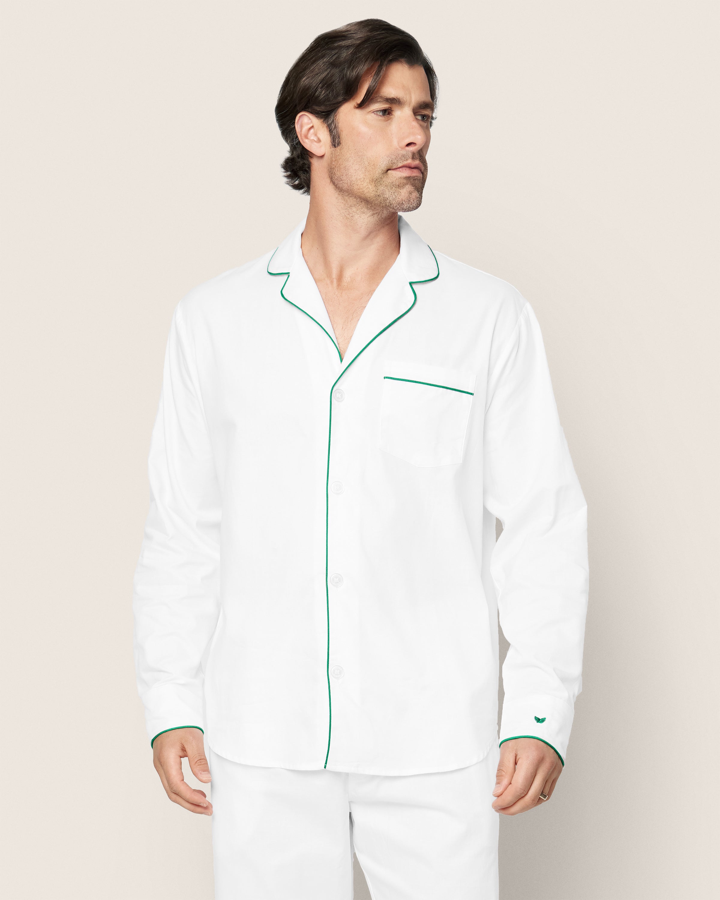 A man models the Petite Plume Mens Twill Pajama Set, featuring a white top with green piping on the collar, cuffs, and pocket. Crafted from premium cotton, his brown hair stands out against a plain, light backdrop.#Color_White with Green Piping