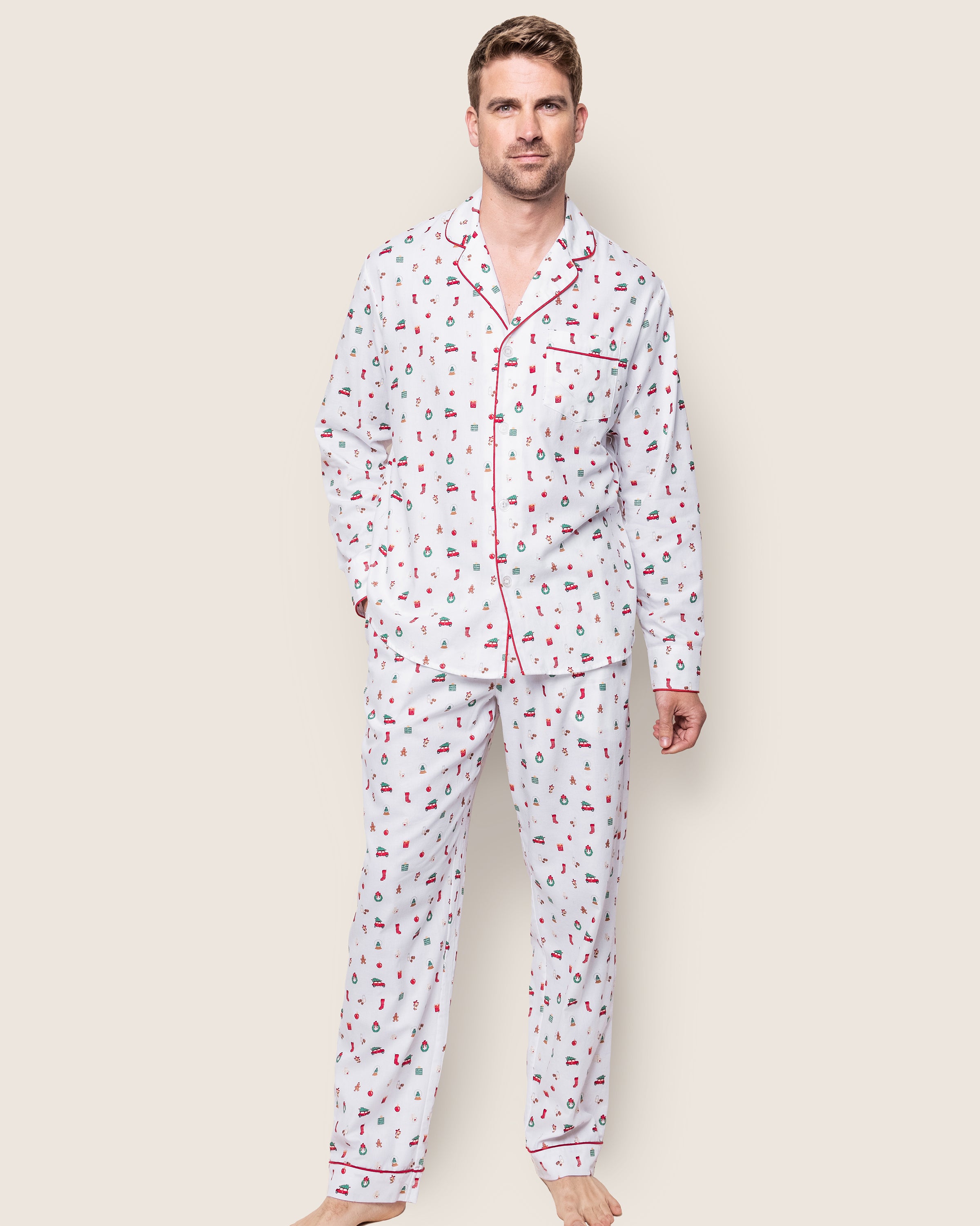 A man stands barefoot in Petite Plumes Mens Twill Pajama Set in Winter Nostalgia, featuring fine-quality cotton with colorful prints and red piping. The set includes a long-sleeve button-up top and matching pants for holiday nights on a plain light background.