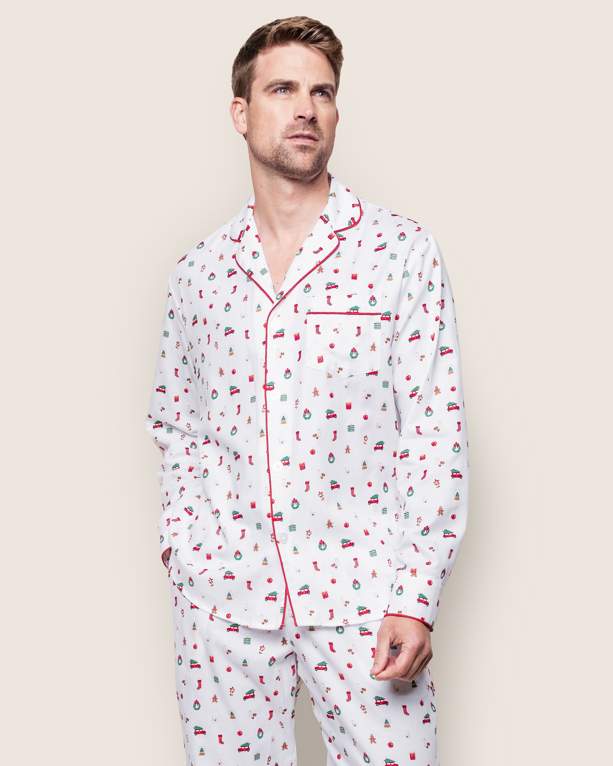 Dressed in the Mens Twill Pajama Set in Winter Nostalgia by Petite Plume, featuring a white top with red piping and a front pocket made from premium cotton, the person looks slightly upward against a light, neutral background, highlighting this festive ensemble.
