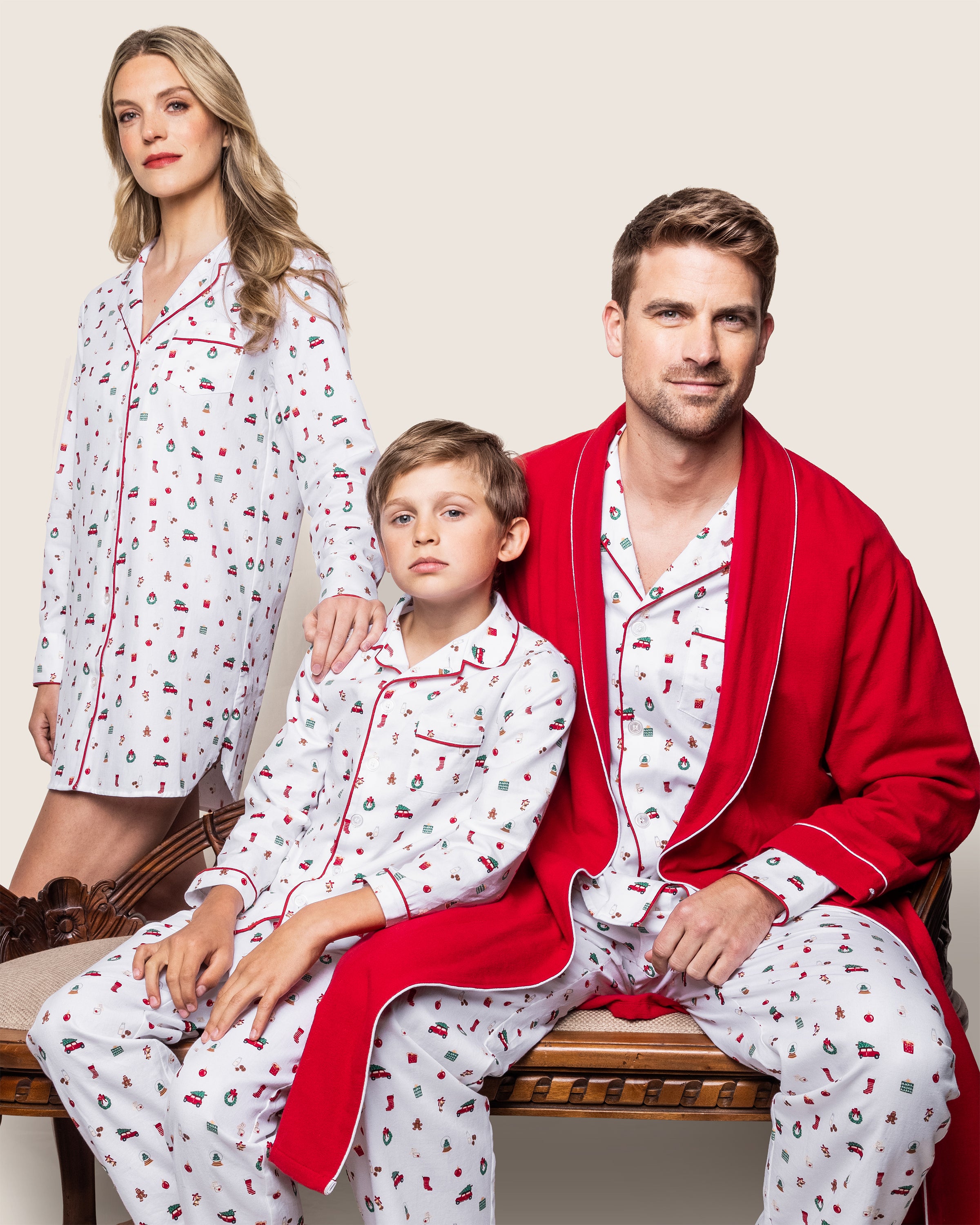 A family, all in matching yarn-dyed cotton pajamas with red designs by Petite Plume, sits together. The man wears a Mens Twill Pajama Set in Winter Nostalgia and a red robe. The woman stands while the rest sit on a bench, blending into the plain background like the gentle chime of Christmas bells.