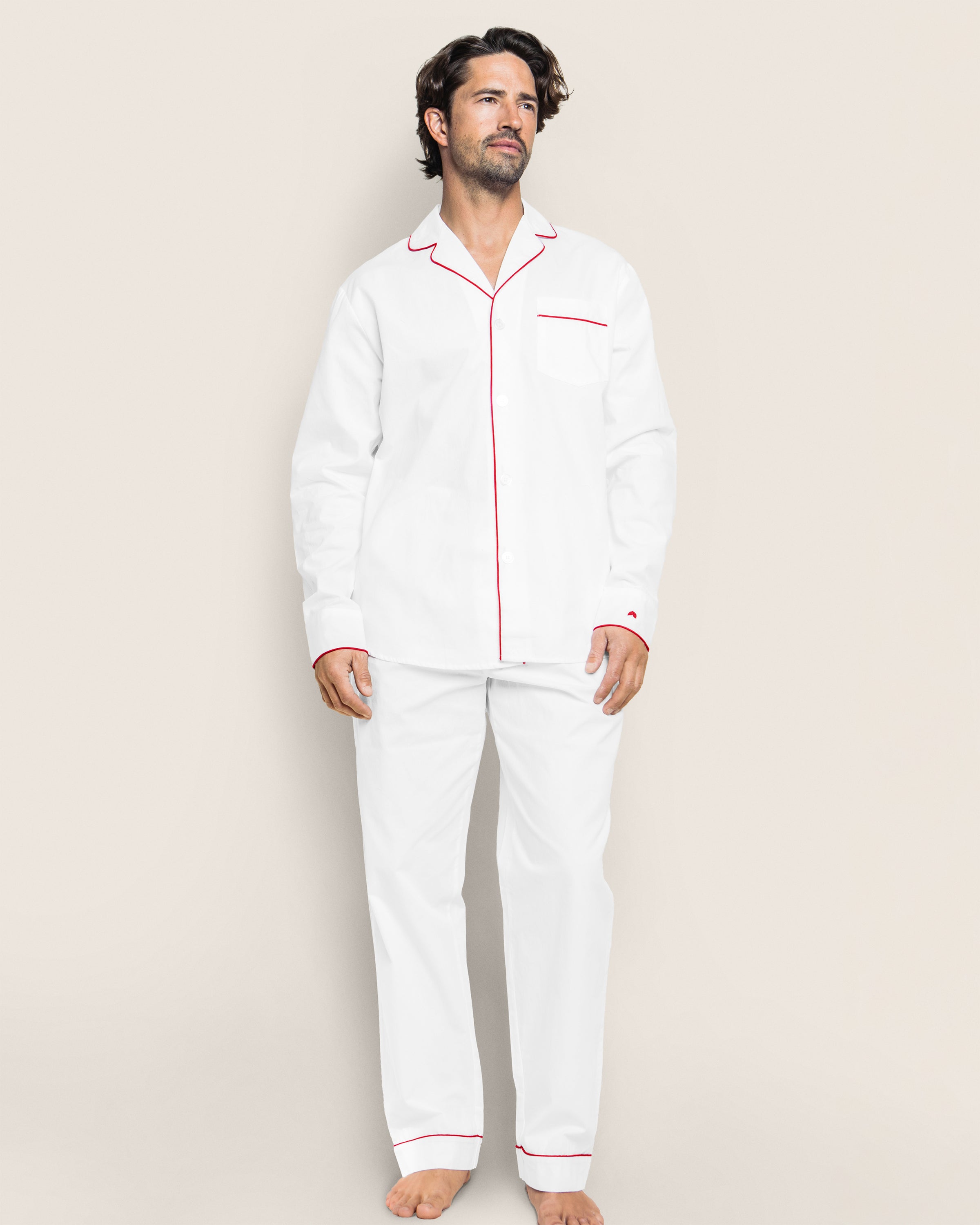 Against a plain light background, a man barefoot and gazing upward embodies elegance in the Petite Plume Mens Twill Pajama Set in White with Red Piping. Made from premium cotton, this luxurious pajama set includes a button-up shirt and matching pants.