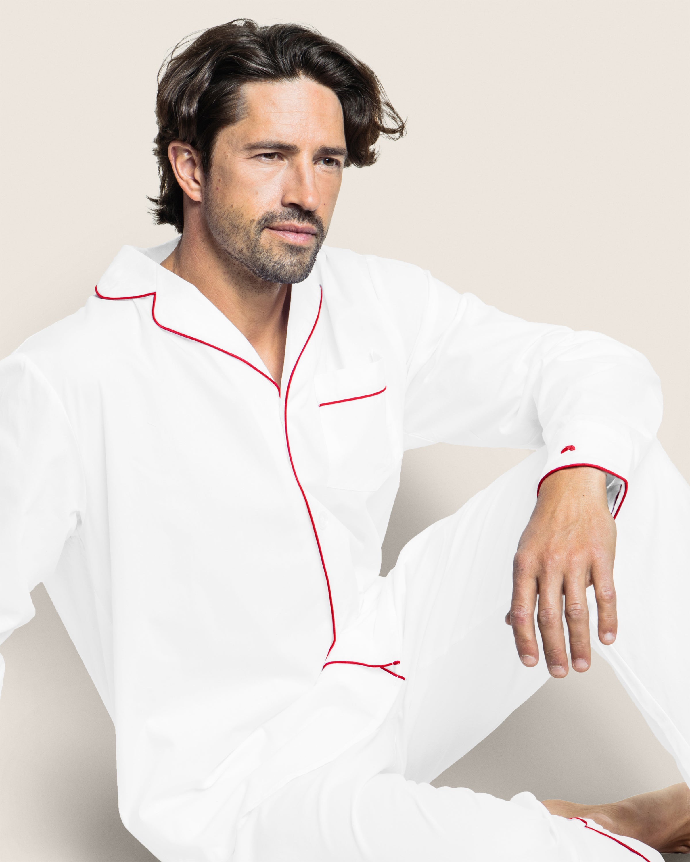 A person with medium-length dark hair and a beard sits in a luxurious Petite Plume Mens Twill Pajama Set in White with Red Piping. They gaze into the distance with a neutral expression against a plain light background.