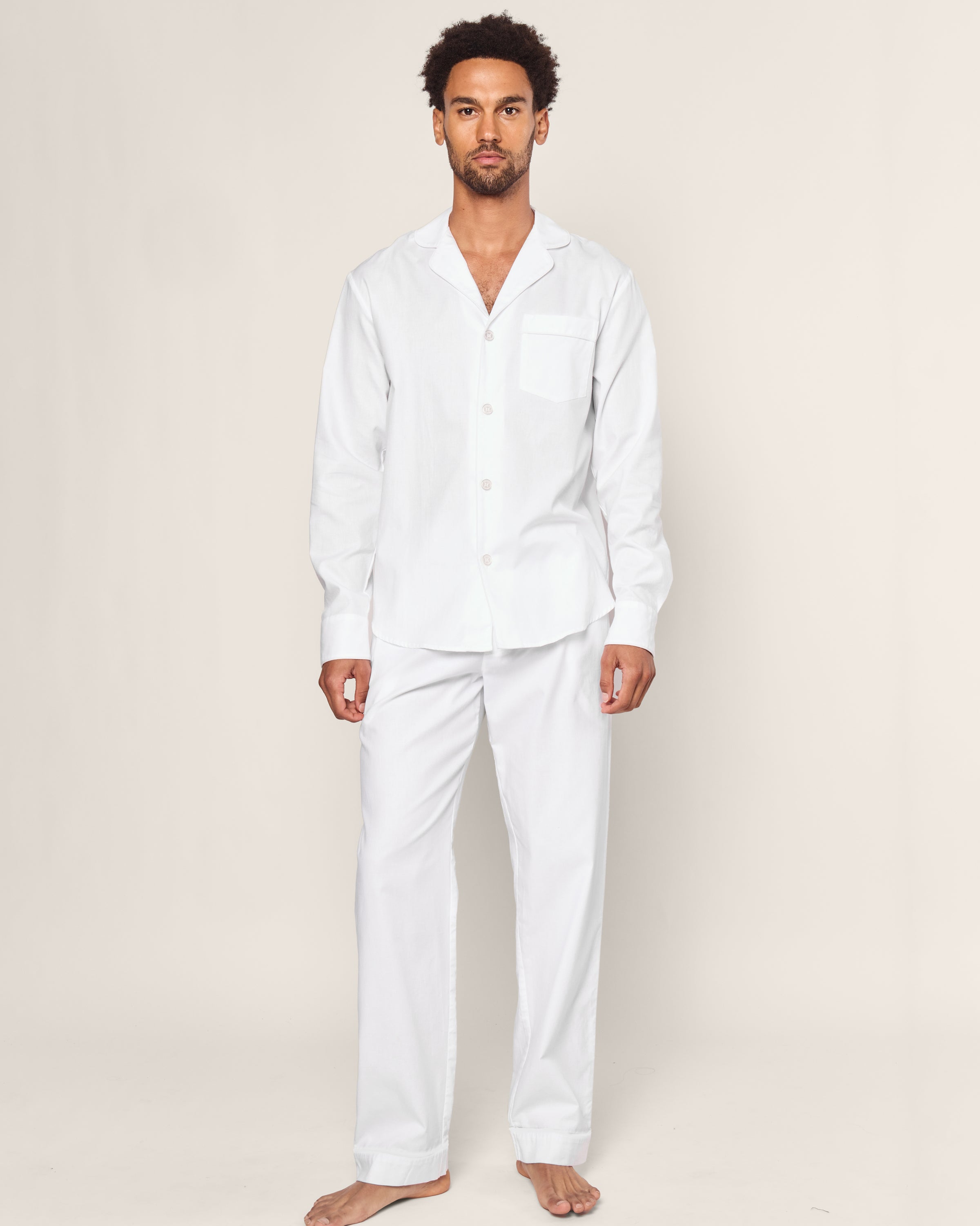 A man in Mens Twill Pajama Set, by Petite Plume, stands barefoot against a neutral background. With short hair and a beard, he wears luxurious cotton sleepwear featuring a long-sleeve button-up shirt with a pocket and matching pants designed with exquisite detail.#Color_White