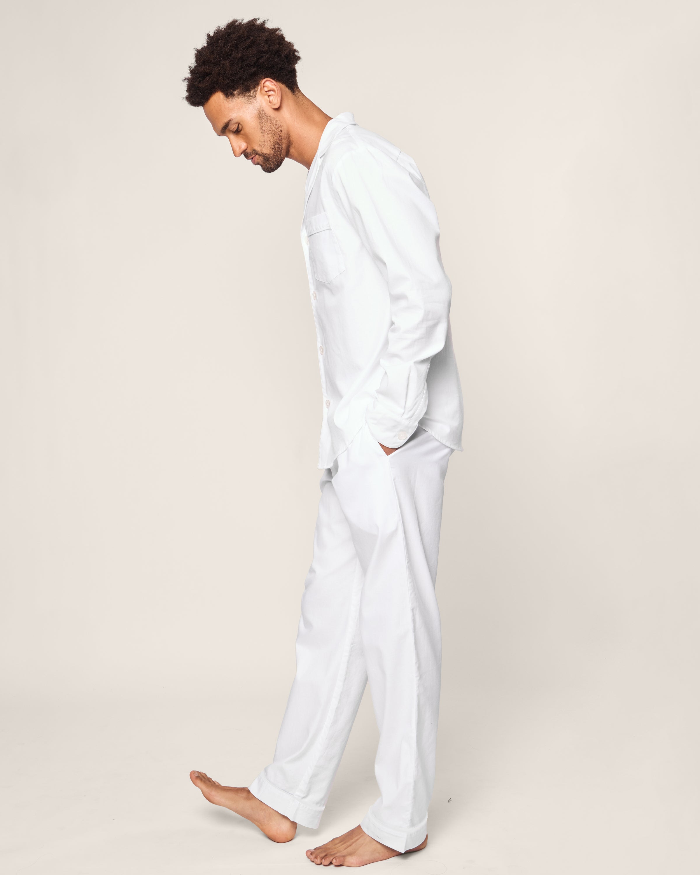 A barefoot person in white pajamas stands against a plain background, looking down with hands in pockets. Theyre wearing the Mens Twill Pajama Set in White with White Piping by Petite Plume, featuring long sleeves and pants for a relaxed and comfortable look.#Color_White