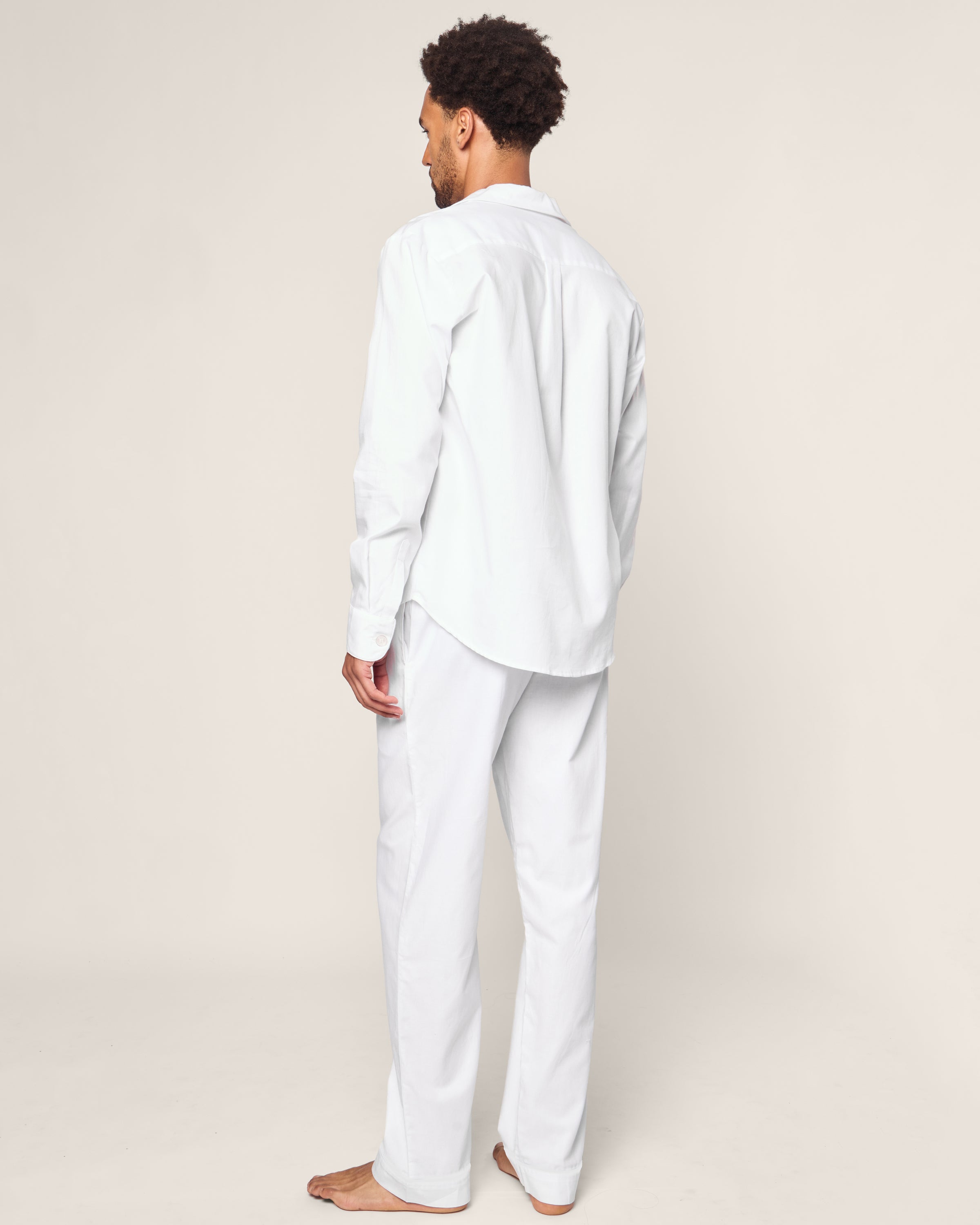 On a plain background, a person stands barefoot in the Mens Twill Pajama Set by Petite Plume, showcasing its luxurious comfort and elegance. The white long-sleeve shirt with matching pants features subtle white piping for a touch of simple sophistication.#Color_White