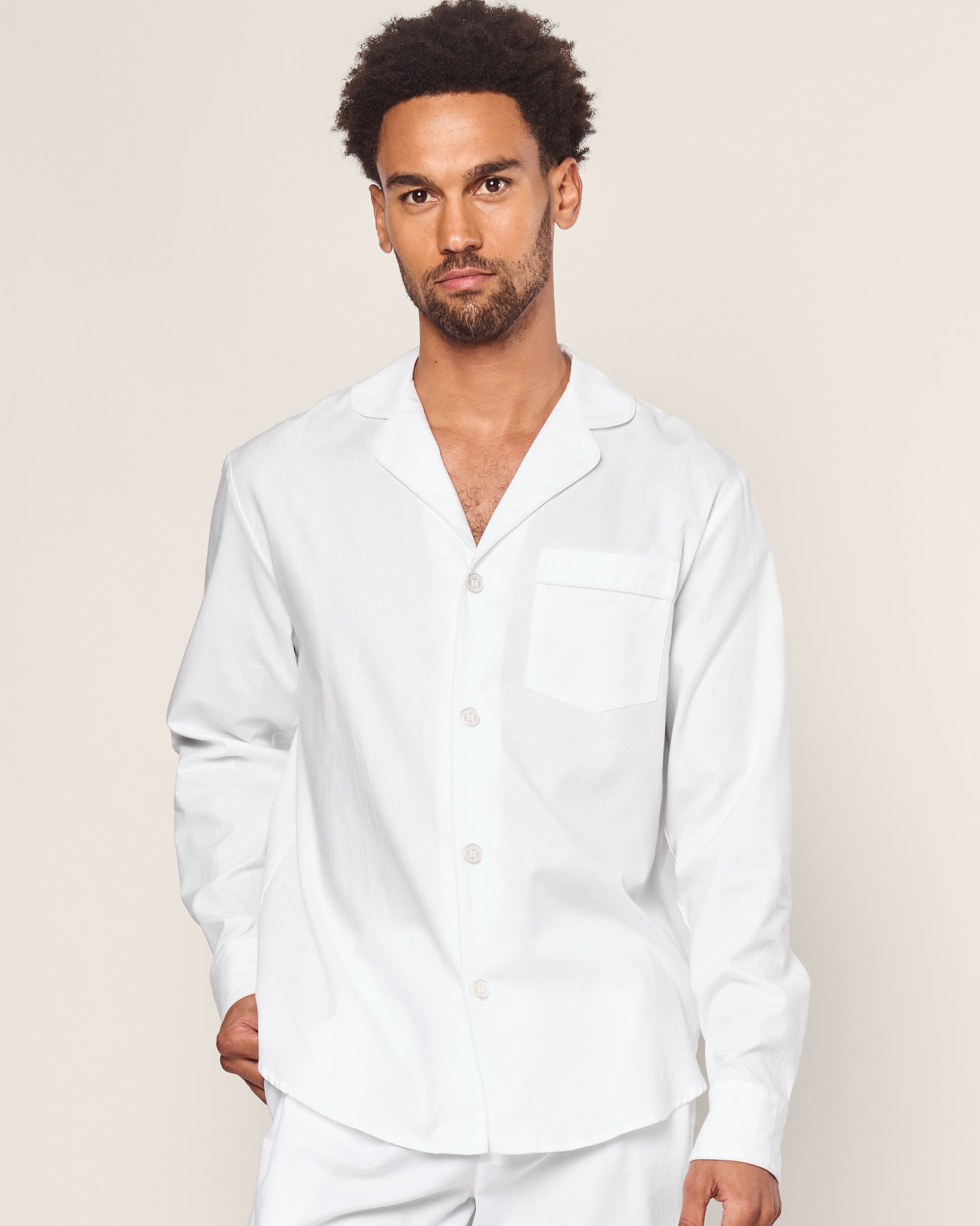A person with short curly hair, wearing a yarn-dyed white button-up shirt with a collar and chest pocket from Petite Plumes Mens Twill Pajama Set in White with White Piping, stands against a plain background, their left hand partially in their pant pocket.#Color_White