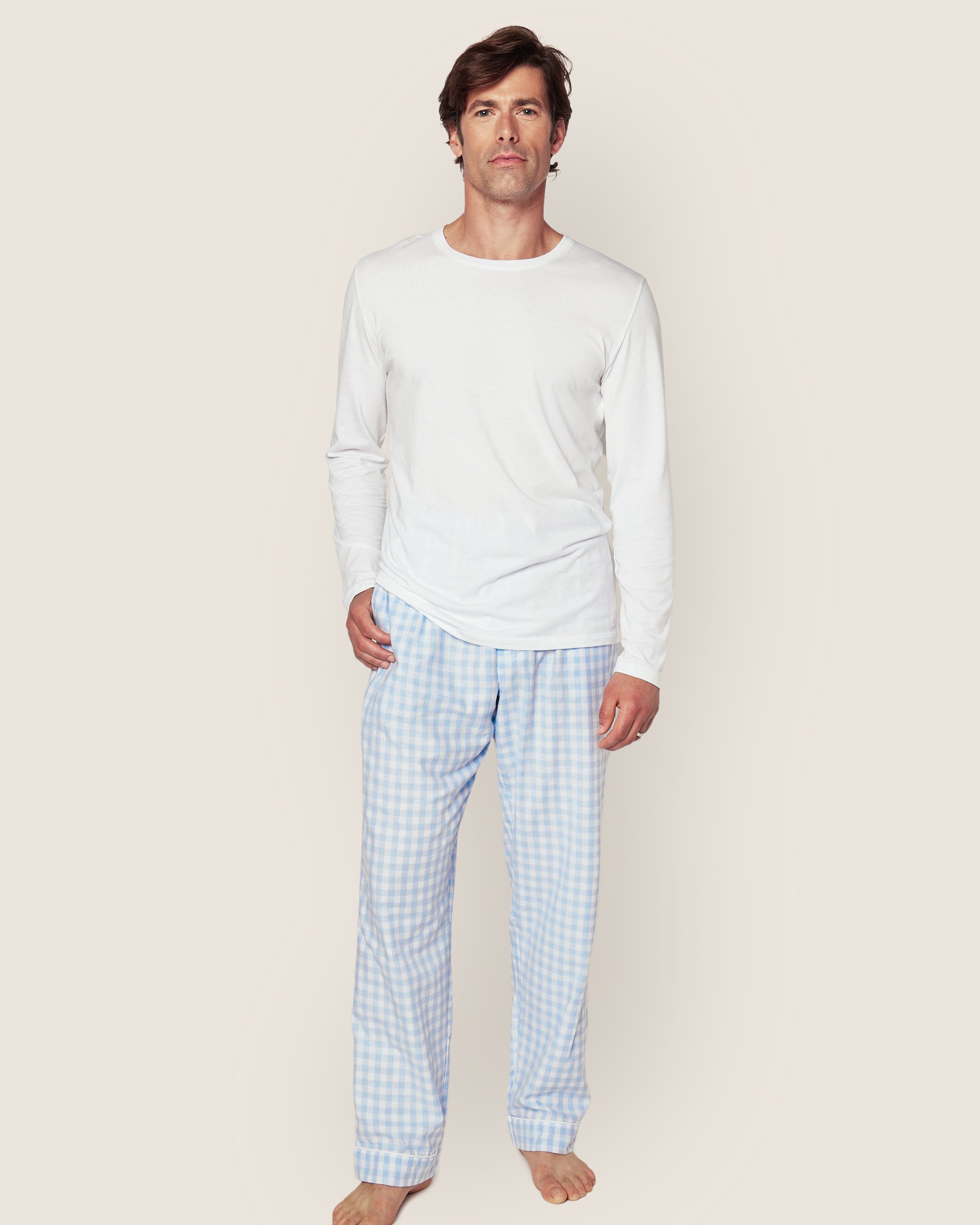 A man stands against a plain background in luxurious sleepwear, wearing a long-sleeved white shirt with Petite Plumes Mens Twill Pants in Light Blue Gingham. He is barefoot and wears a neutral expression while looking directly at the camera.