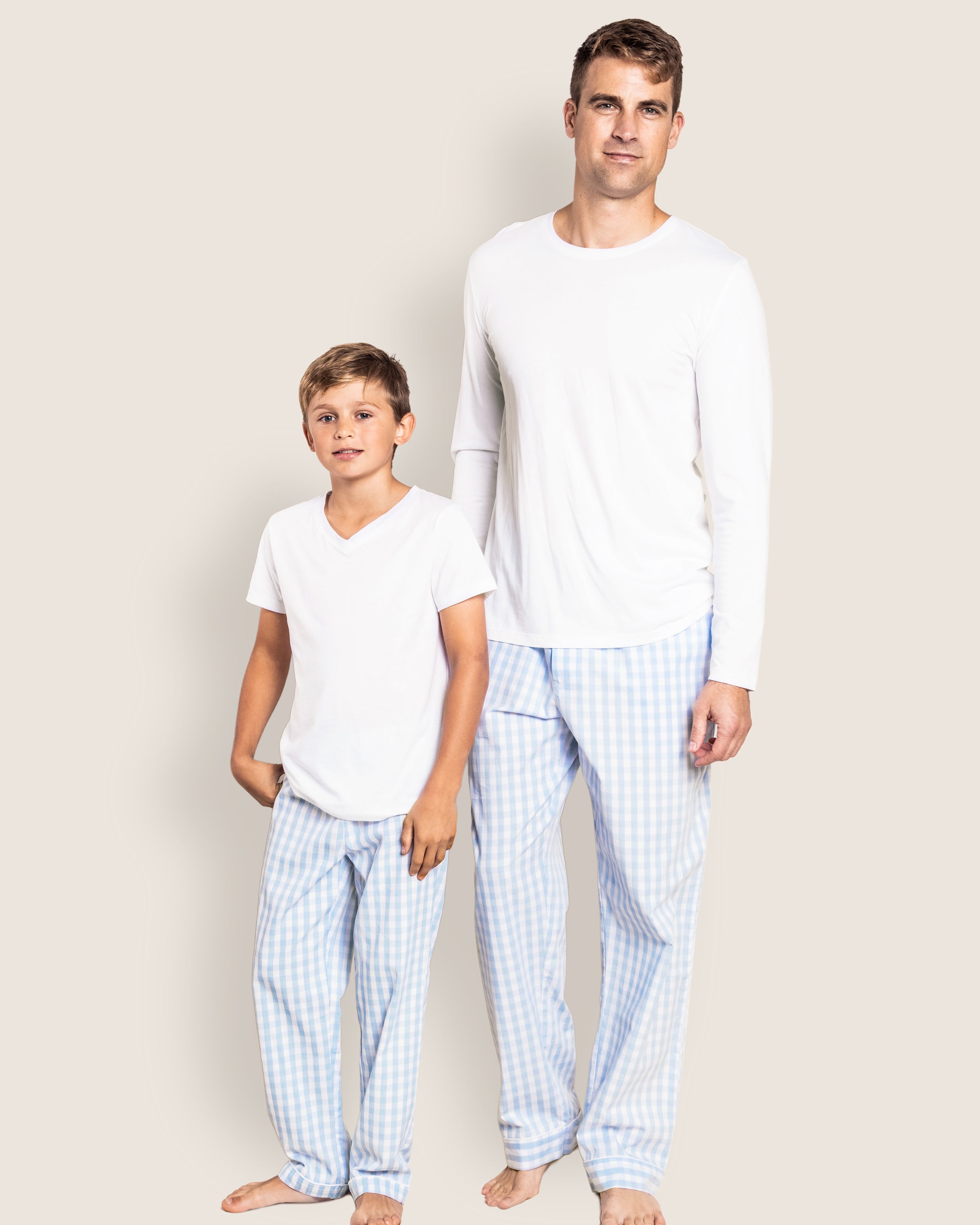 An adult and a child stand barefoot against a plain background, wearing Petite Plume sleepwear: white tops with Mens Twill Pants in Light Blue Gingham.