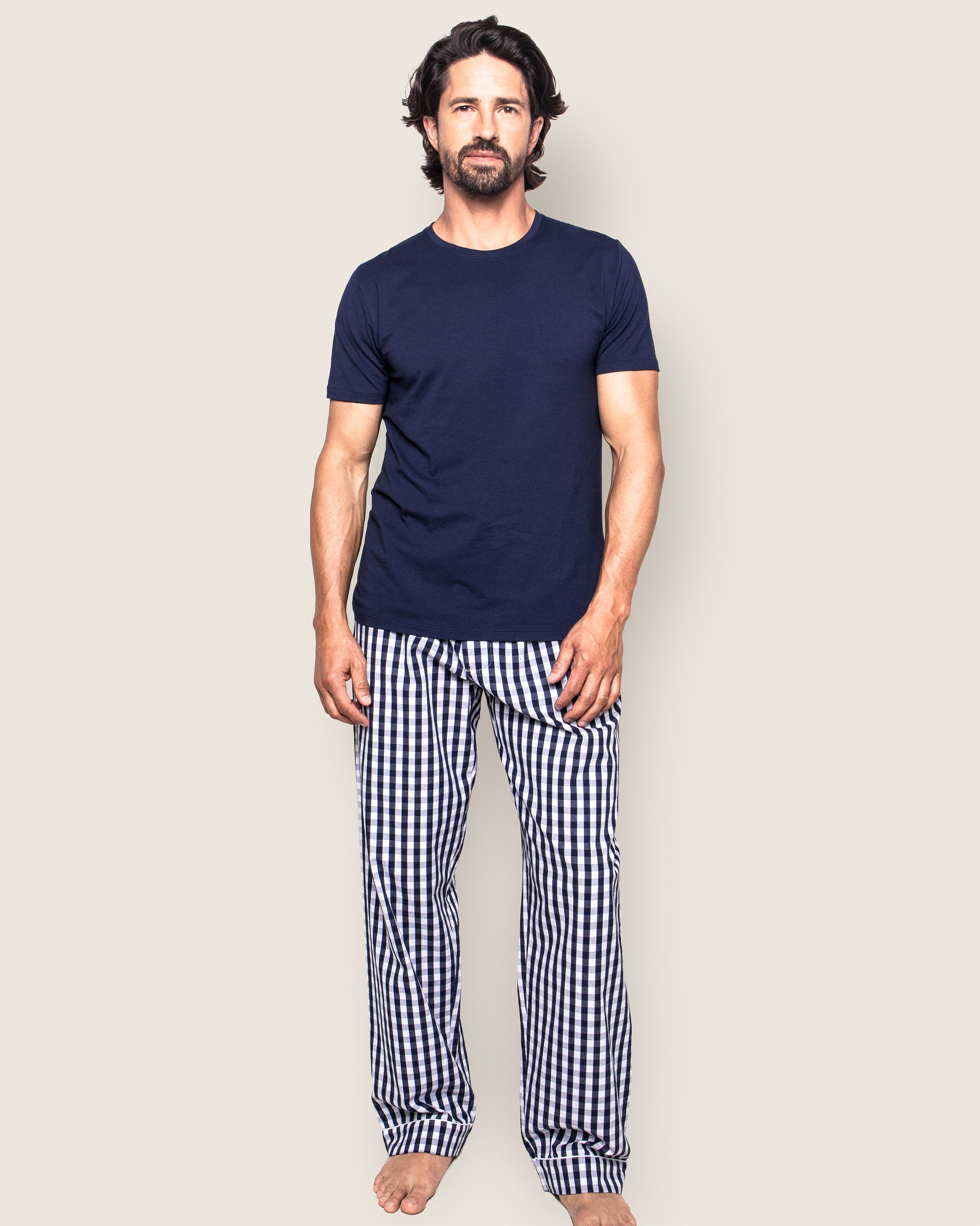A man with dark hair and a beard stands barefoot against a neutral background, wearing a navy blue t-shirt and Petite Plumes Mens Twill Pajama Pant in Navy Gingham.