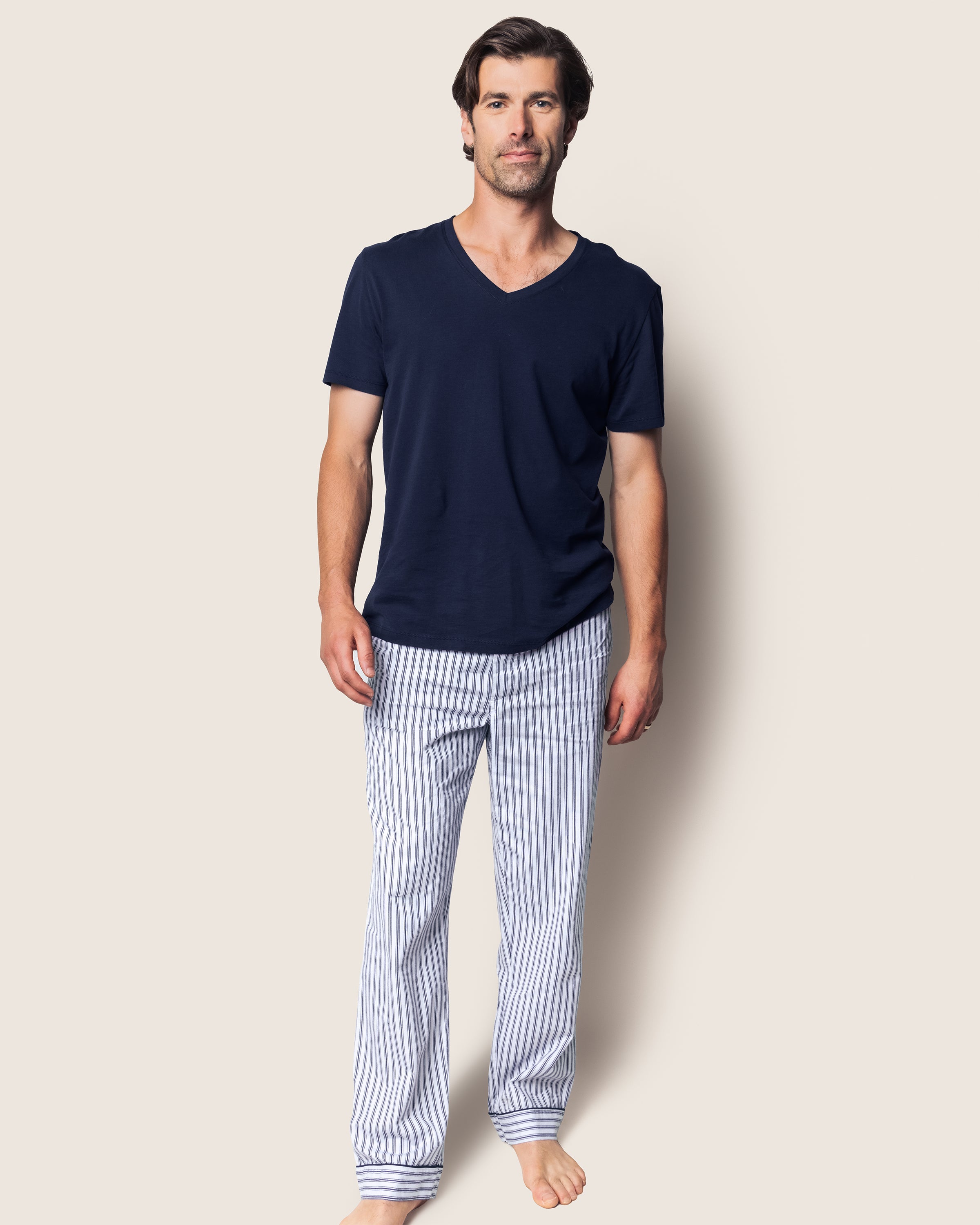 A person stands against a plain background, barefoot and relaxed, wearing Petite Plumes luxurious Mens Twill Pajama Pants in Navy French Ticking and a navy V-neck T-shirt, gazing directly at the camera.