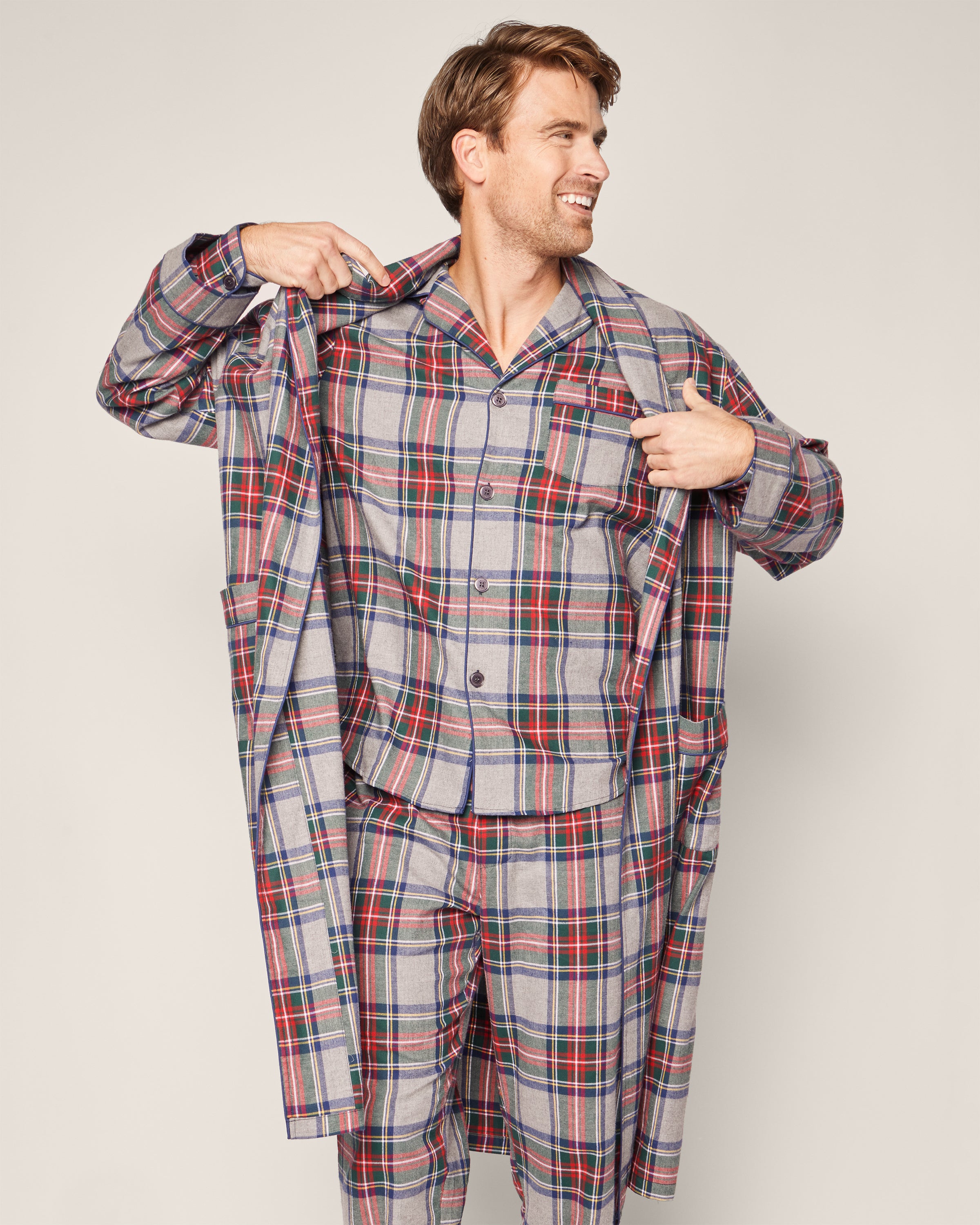 A man smiles while holding open his Petite Plume Mens Brushed Cotton Robe in Westminster Tartan, revealing matching plaid pajamas. He stands against a plain, light background, embodying luxury sleepwear.