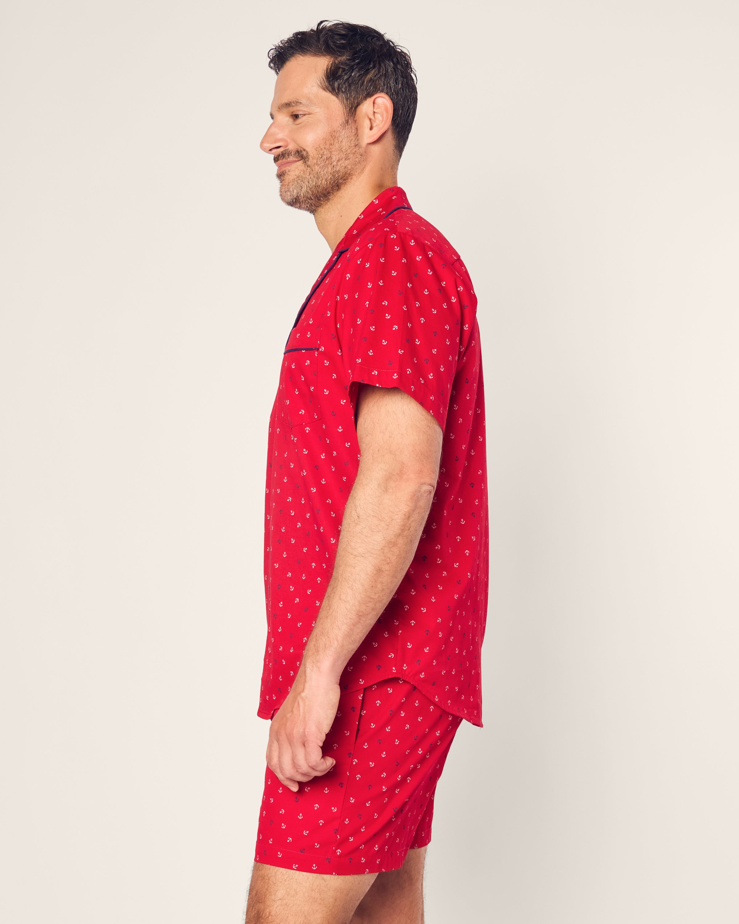 A man stands in profile wearing Petite Plumes Mens Twill Short Sleeve Short Set Anchors Away, featuring a red shirt and matching shorts with an Anchors Away print. The shirt has short sleeves with black piping, and the plain background emphasizes the vibrant colors of premium cotton.