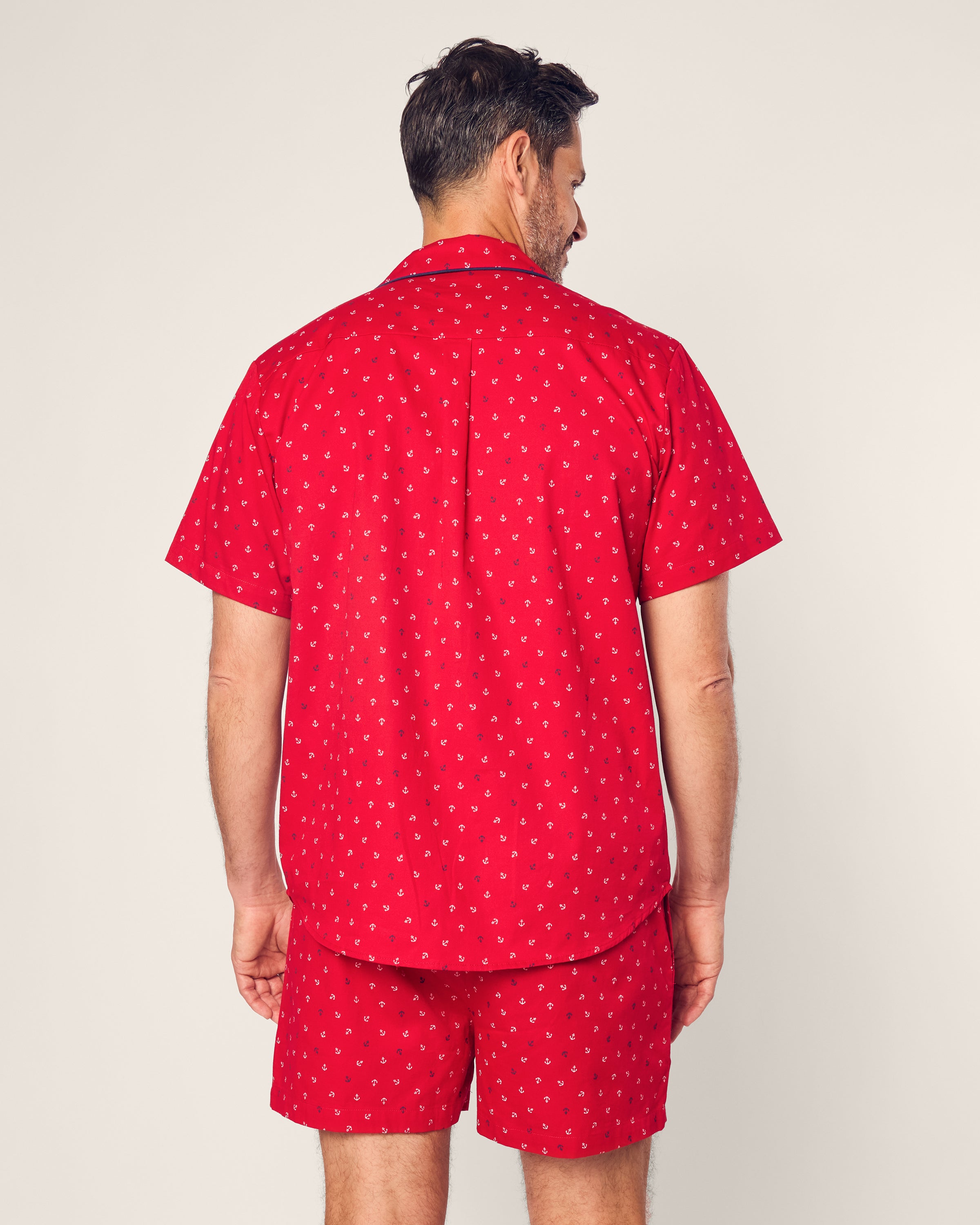A man with short, dark hair stands against an off-white background wearing Petite Plumes Mens Twill Short Sleeve Short Set in Anchors Away print, exuding relaxed sophistication in classic red pajamas crafted from high-quality cotton.
