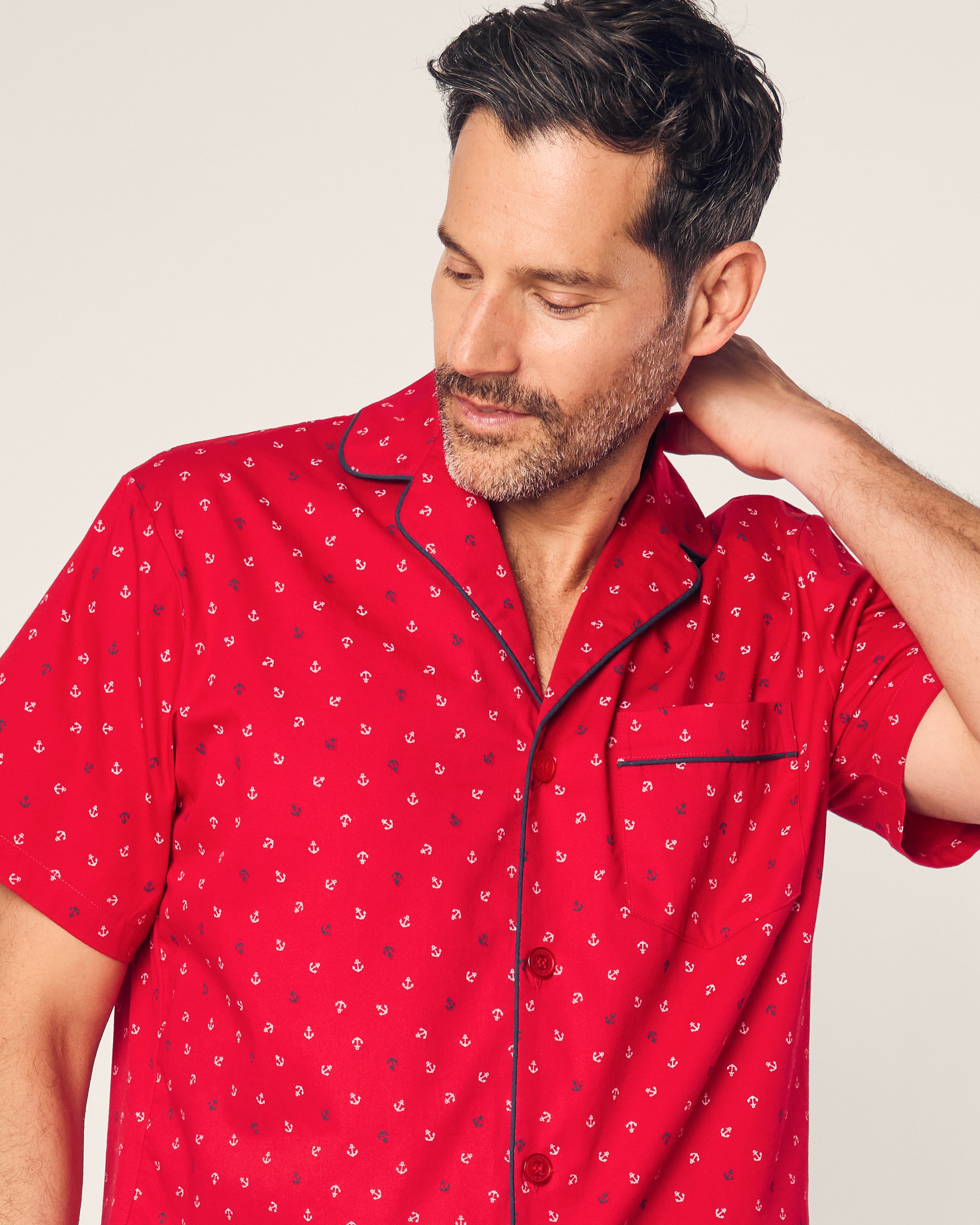 A man with short dark hair and facial hair wears a Petite Plume Mens Twill Short Sleeve Short Set Anchors Away in red. Hes looking down, touching the back of his neck with his left hand against a plain background.