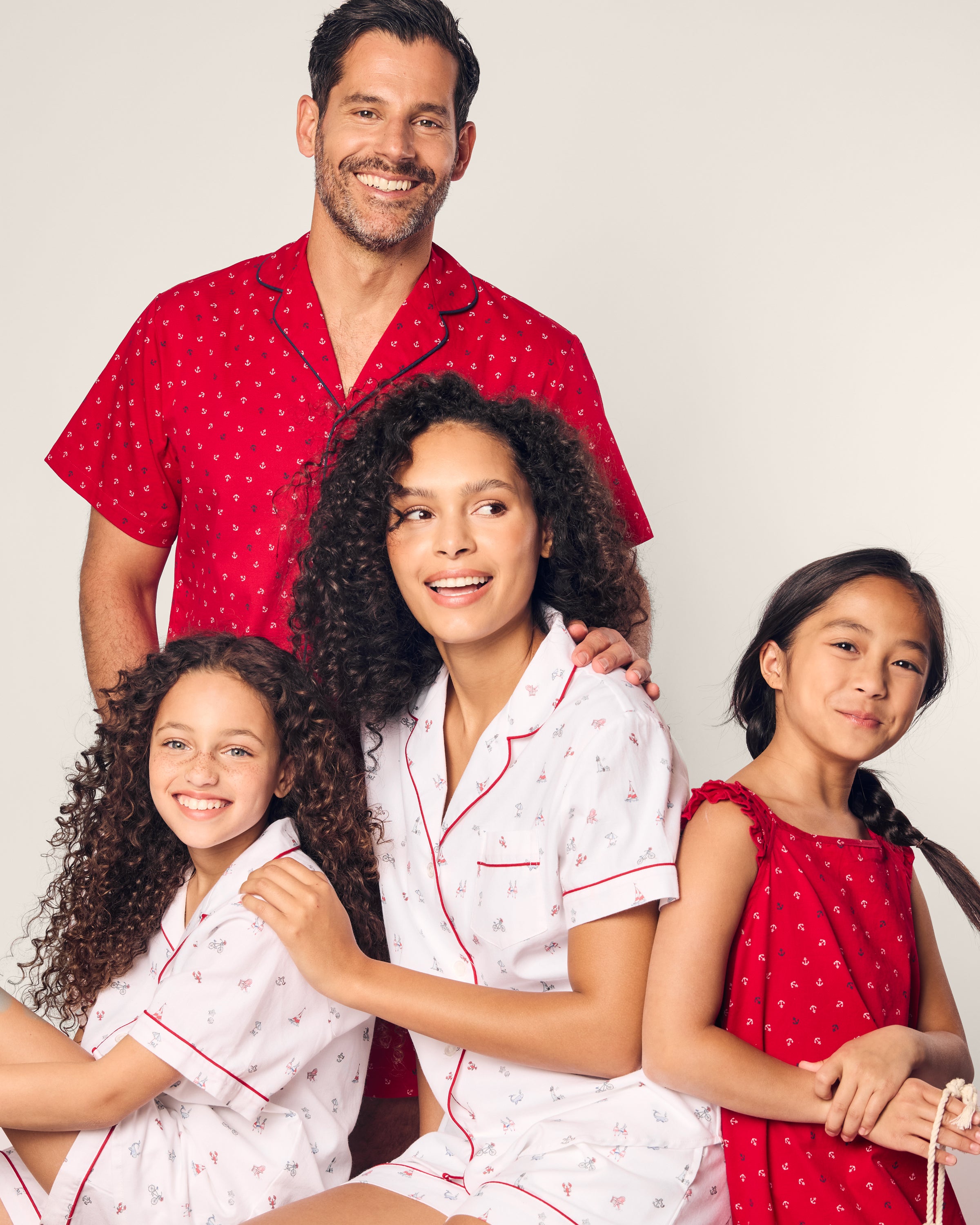 In a cheerful family photo against a light background, the father is dressed in Petite Plumes Mens Twill Short Sleeve Short Set Anchors Away. The mother and young daughter wear Classic Short Sets with the same print, while the older girl stands out in a charming red dress. All are smiling and looking in different directions.