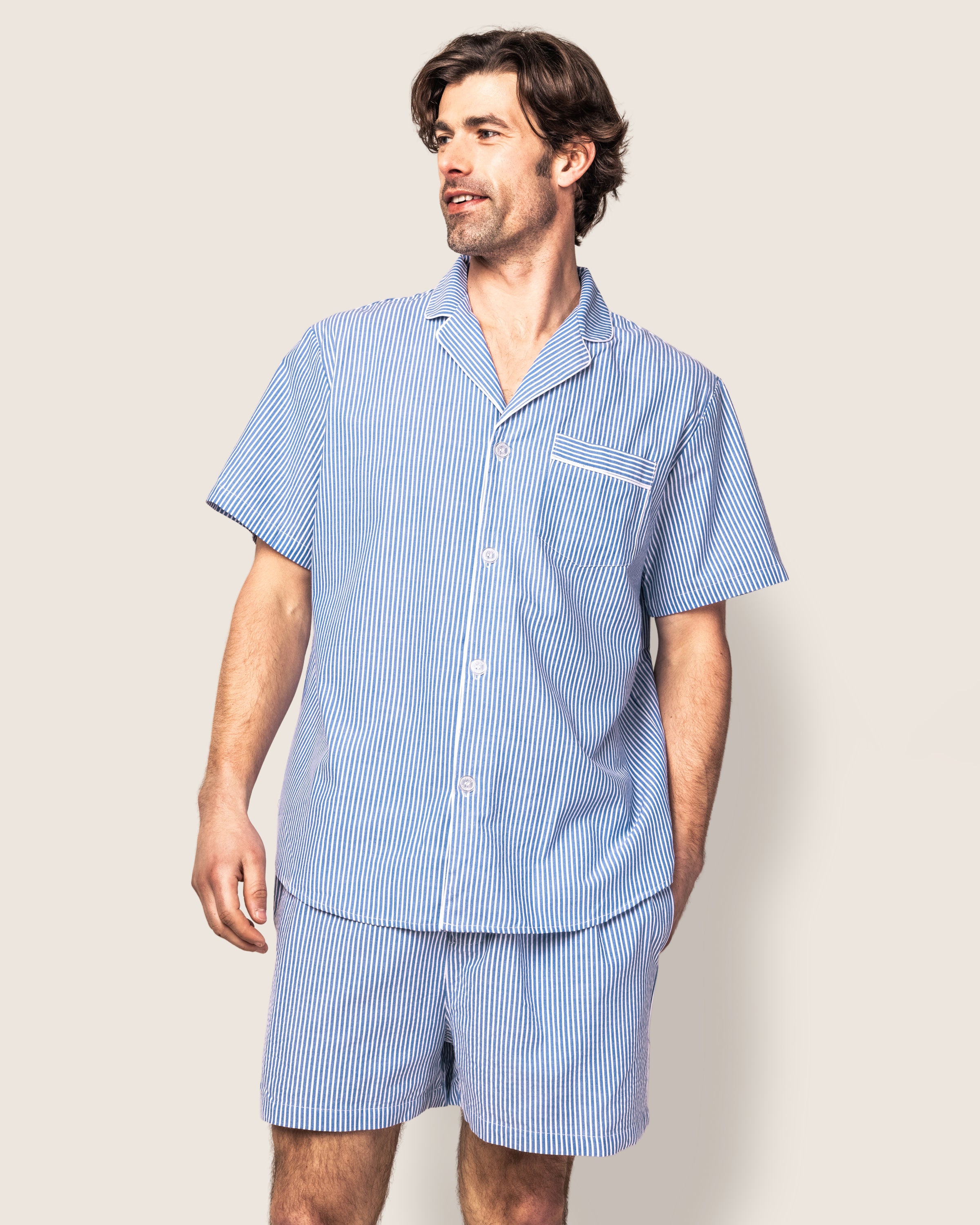A man models the Mens Twill Pajama Short Set in French Blue Seersucker by Petite Plume. This luxurious set, crafted from 100% cotton sleepwear, features a button-up shirt with a small breast pocket and matching shorts against a plain background.