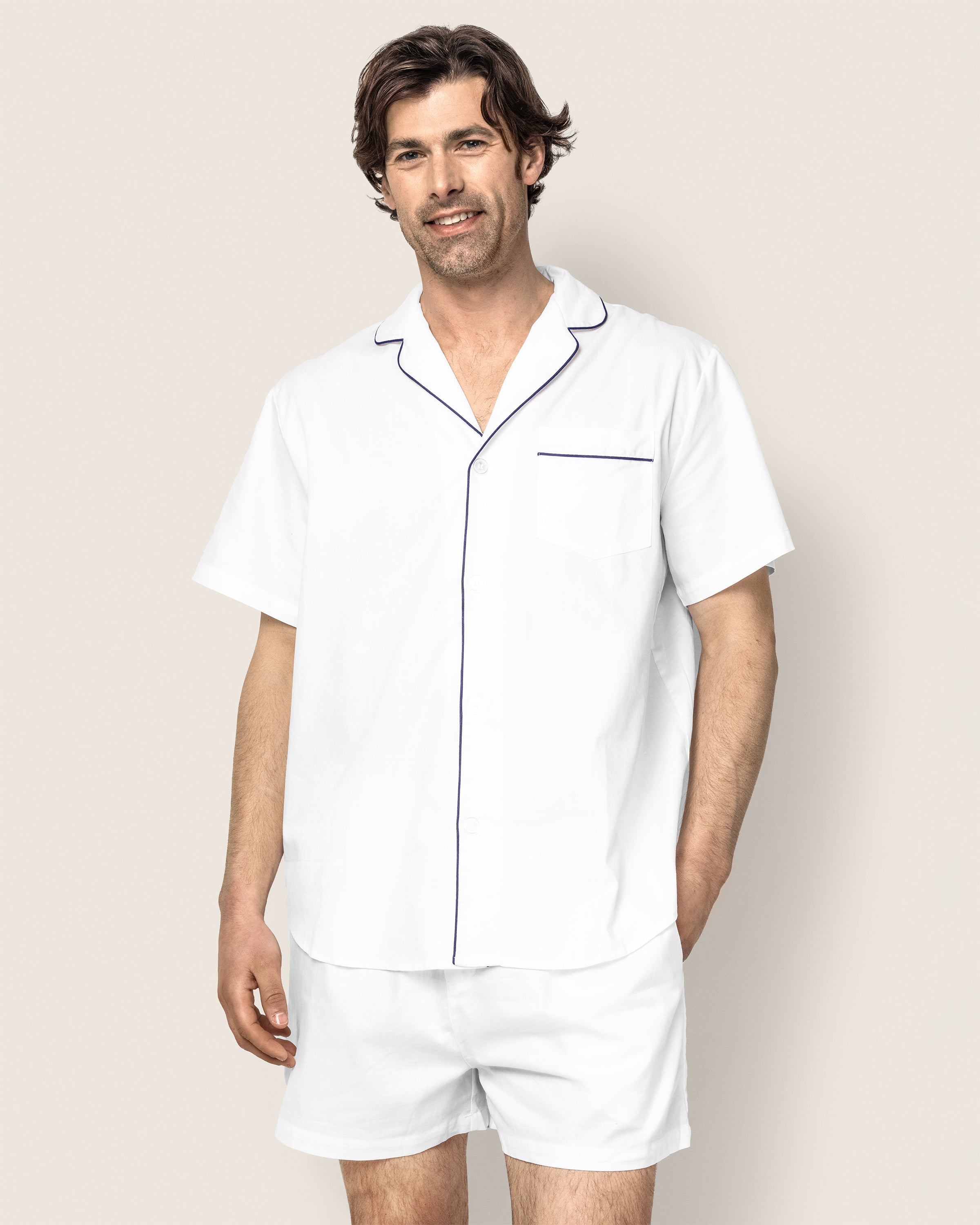 A man smiles in Petite Plumes Mens Twill Pajama Short Set, showcasing 100% cotton comfort. The white ensemble features navy piping, a button-up shirt with a pocket, and matching shorts. Standing against a beige background, he exudes relaxed sophistication.