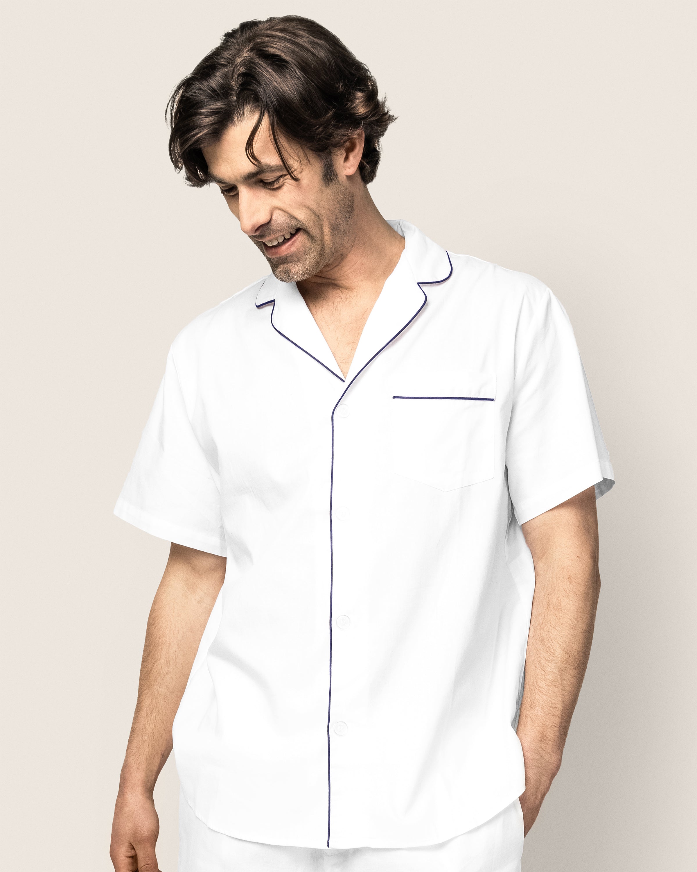 A man wearing Petite Plumes Mens Twill Pajama Short Set in white with navy piping, looking down and smiling against a plain background, exuding the comfort of luxury sleepwear.